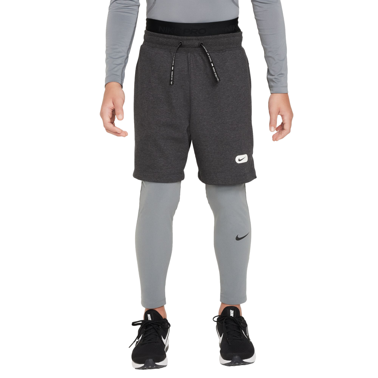 A person wearing grey Nike athletic wear, including fleece shorts over compression leggings and black sneakers, against a solid black background.