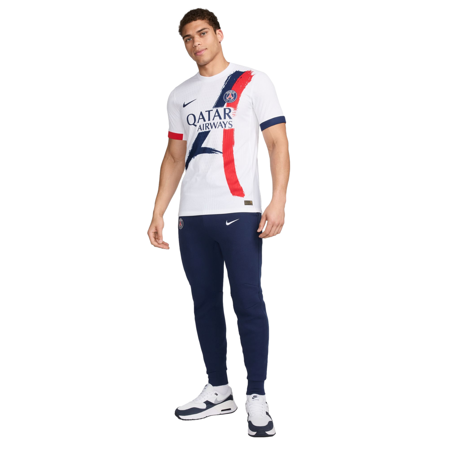 A male model wearing a white Paris Saint-Germain jersey with blue and red graphic details, navy Nike joggers, and white sneakers against a white background.