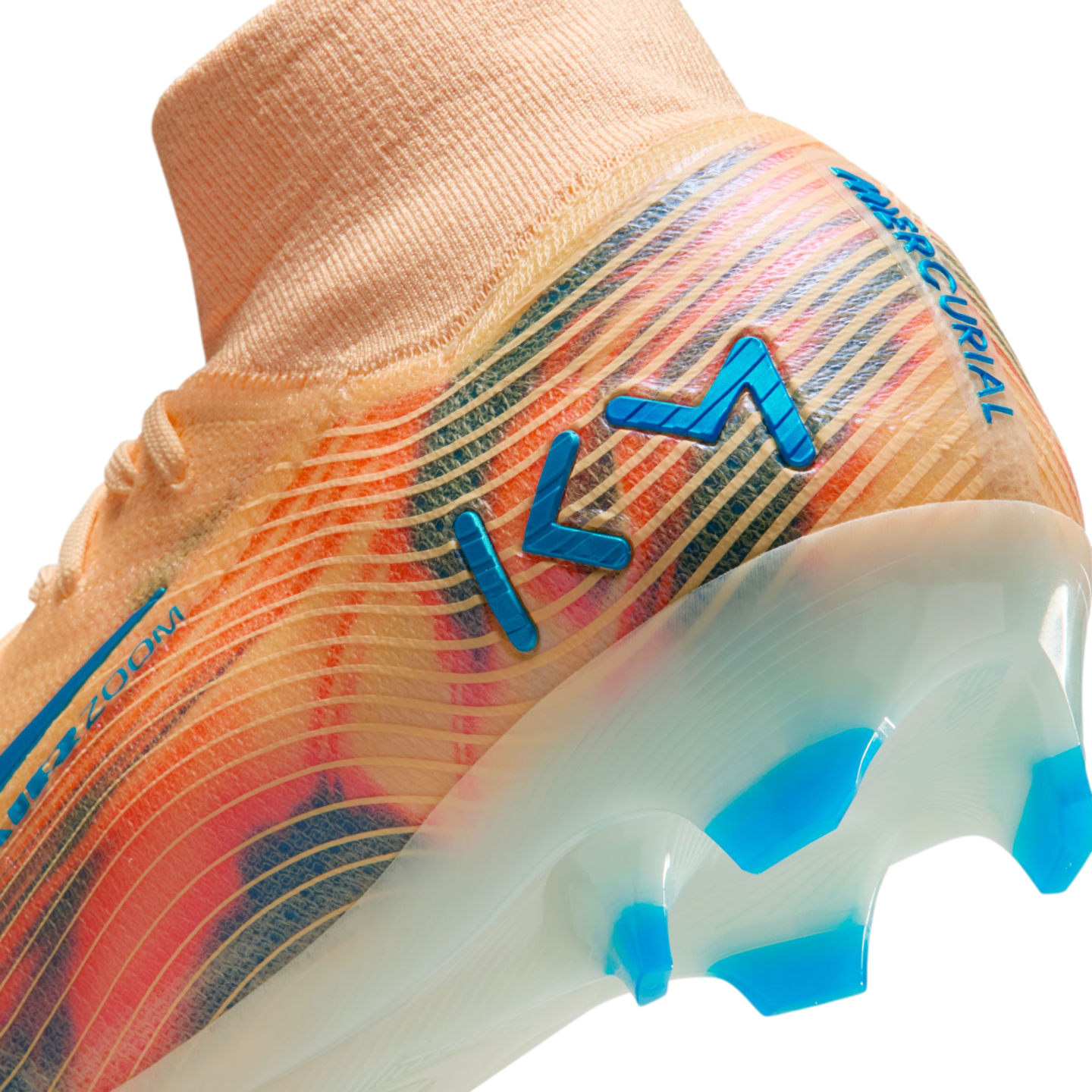 Close-up of a Nike Mercurial soccer cleat in orange with blue and pink accents, featuring a blue "KM" logo and a translucent sole with blue studs.
