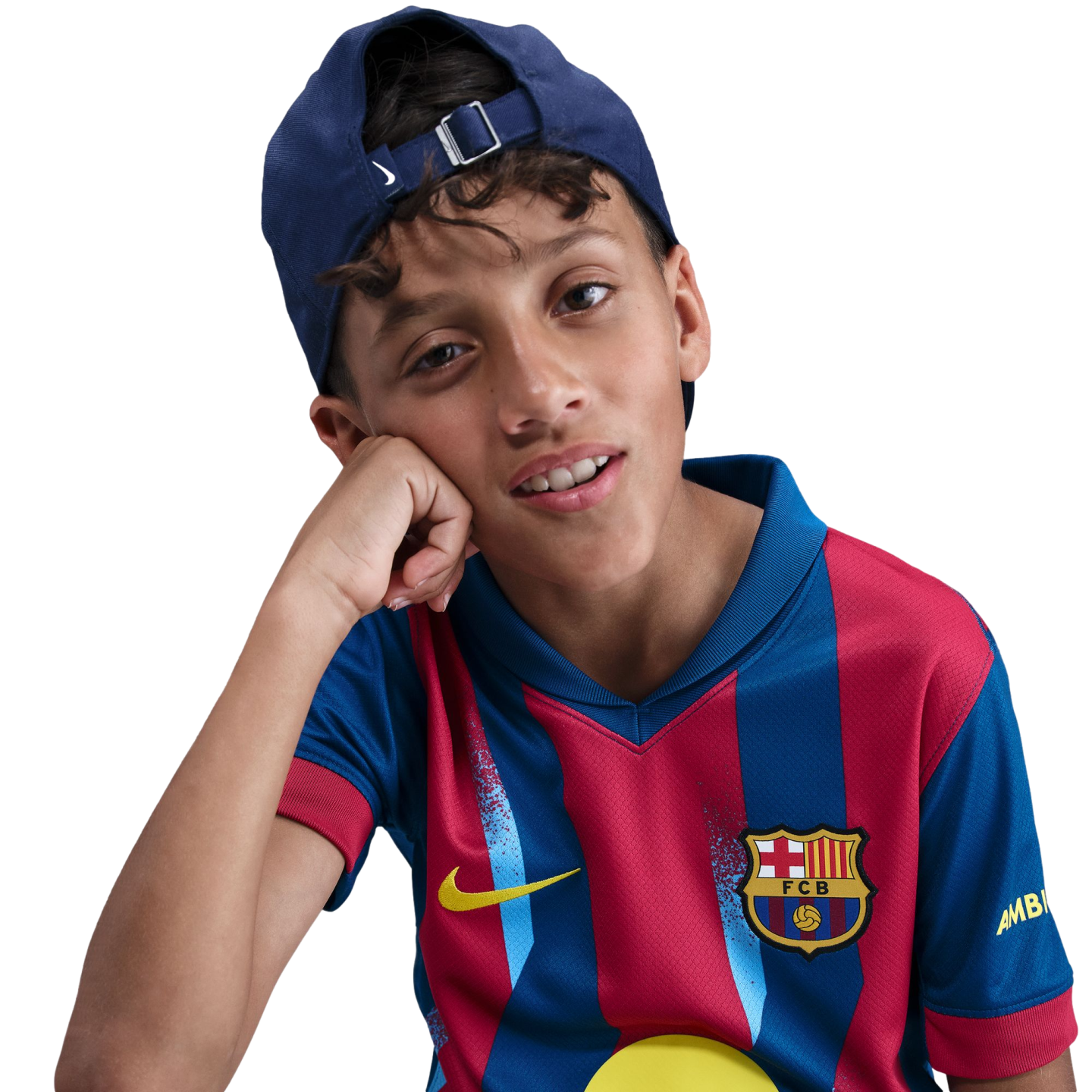 Portrait of a young boy wearing an FC Barcelona jersey and a backwards blue cap, resting his head on his hand against a black background.
