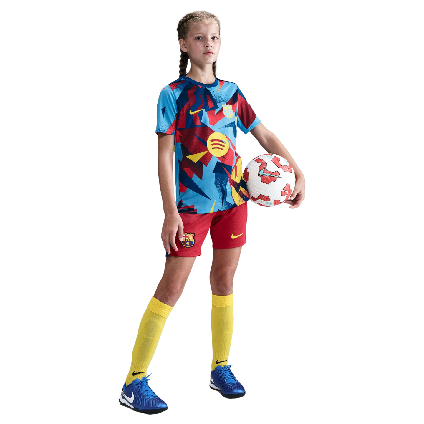 A young girl wearing a patterned soccer jersey, red shorts, and yellow socks stands against a black background, holding a soccer ball under her arm.