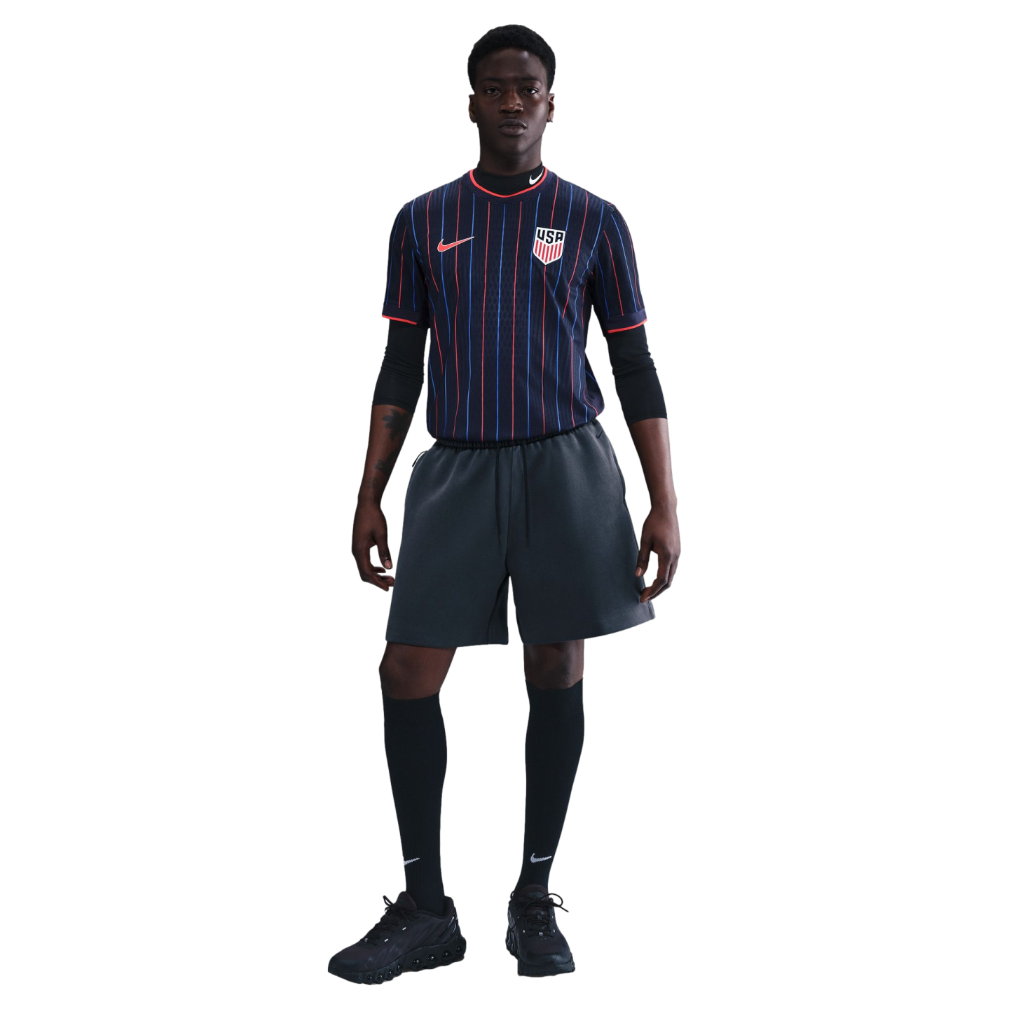 A man models a navy blue USA soccer jersey with red and blue pinstripes, charcoal gray shorts, black knee-high socks, and black sneakers.