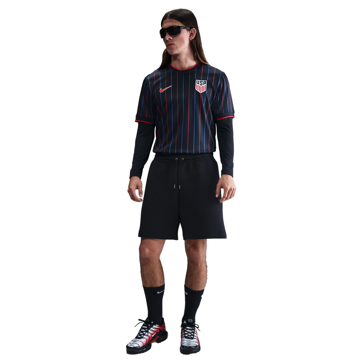 A person with long hair in sunglasses wearing a black pinstriped USA soccer jersey, black shorts, and Nike sneakers against a white background.
