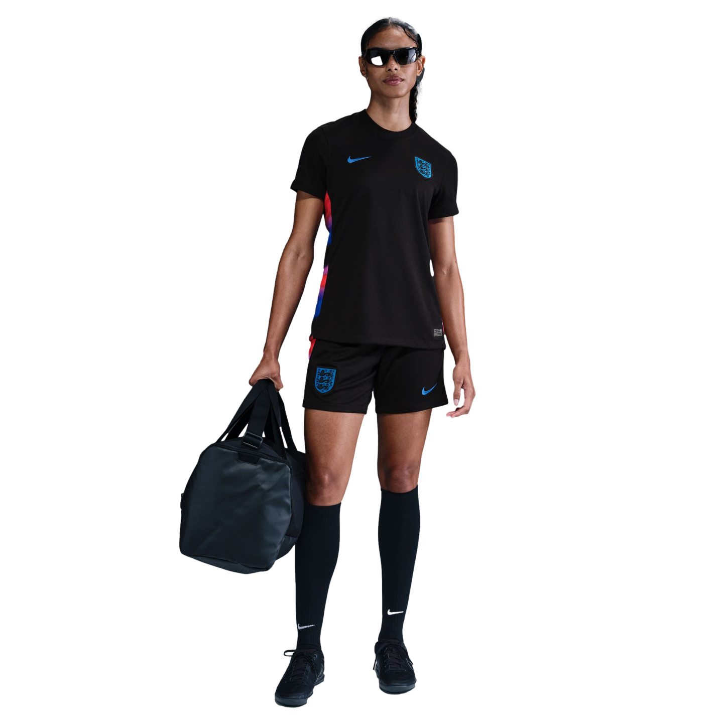 A woman standing in a black Nike England soccer kit and sunglasses, holding a black duffel bag.