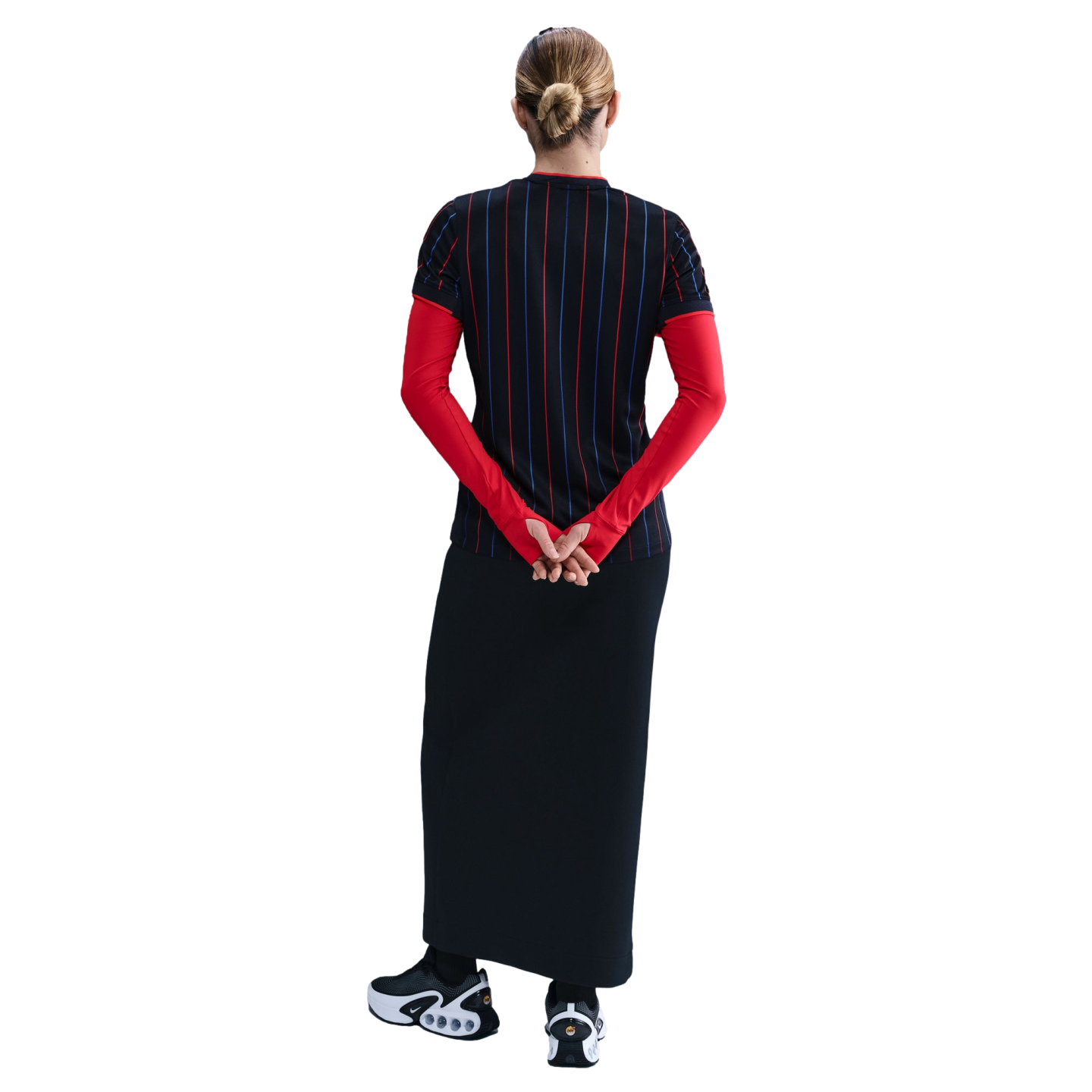 Back view of a person with a bun wearing a black pinstriped top over long red sleeves, a long black skirt, and black and white Nike sneakers.