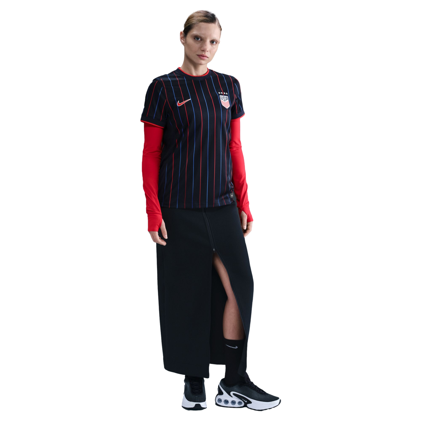 A woman in a striped black USA soccer jersey, red undershirt, long black skirt, and black sneakers, standing against a white background.