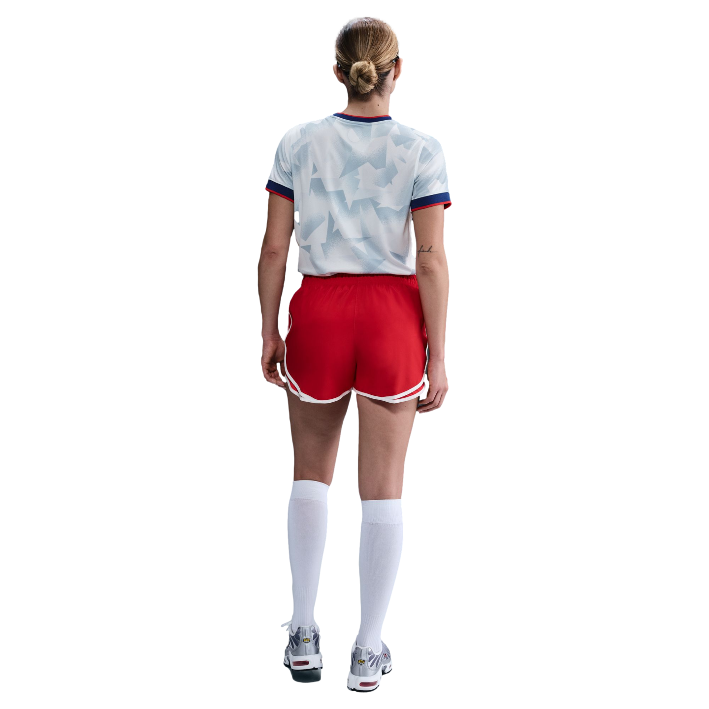 Back view of a person wearing a patterned grey jersey, red athletic shorts, knee-high white socks, and silver sneakers against a white background.
