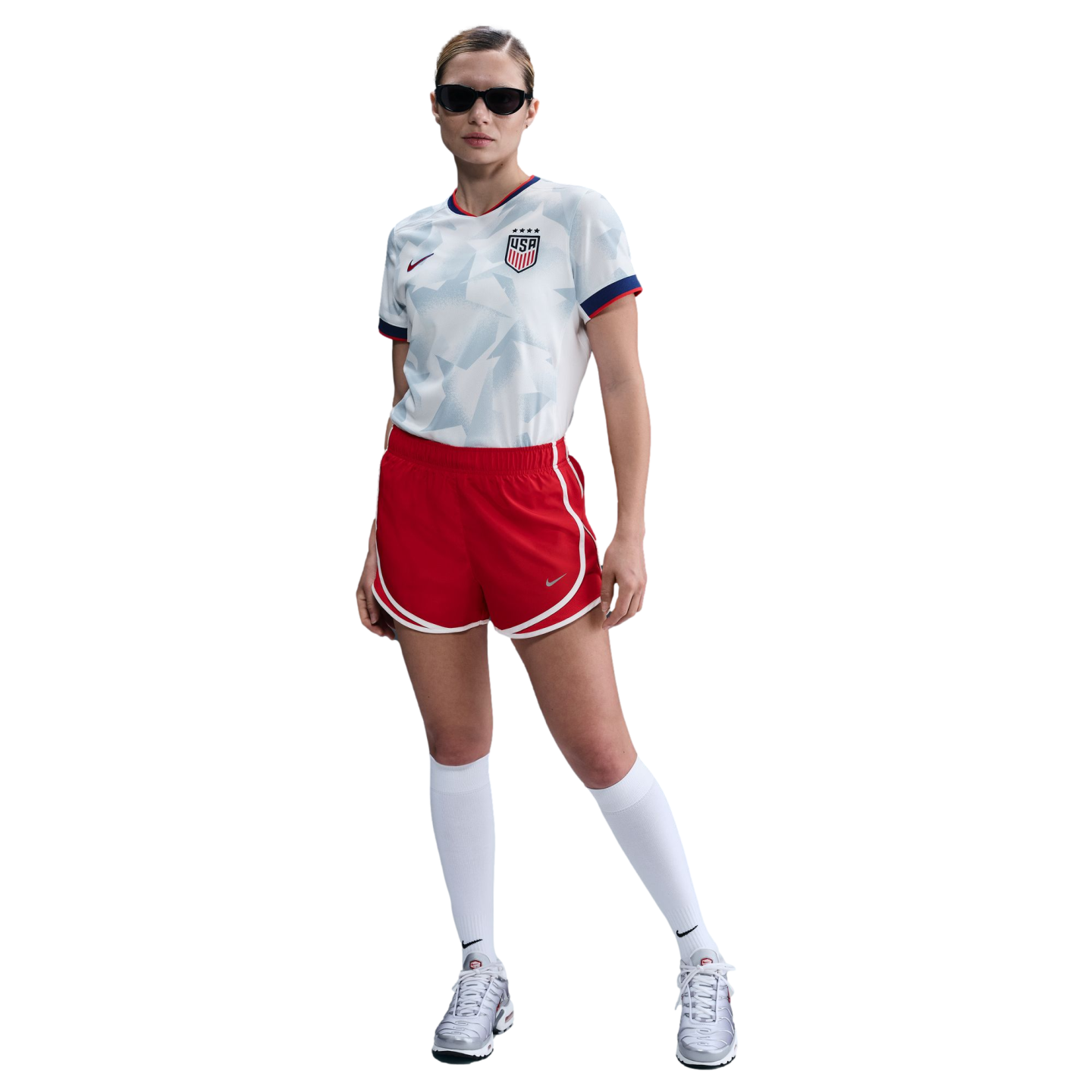 Woman in a white USA soccer jersey, red shorts, white socks, silver sneakers, and black sunglasses.