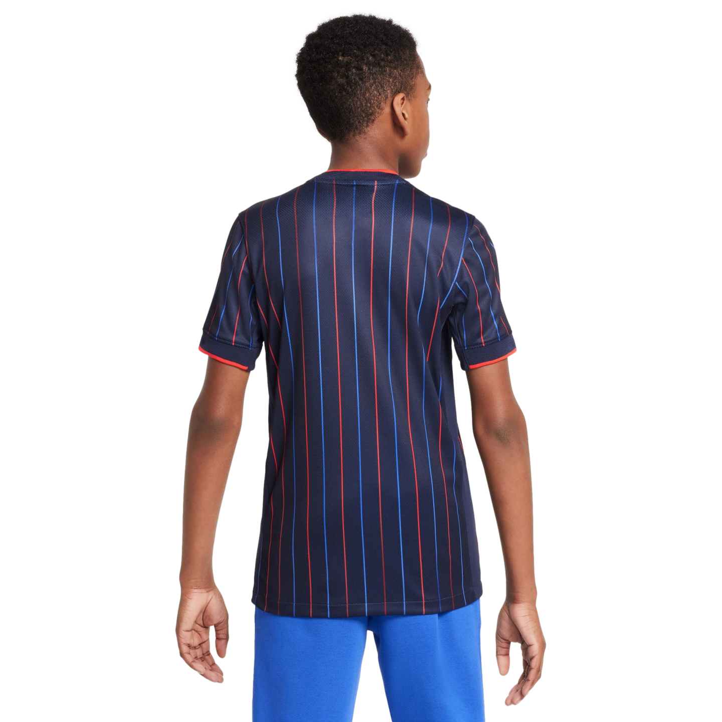 Rear view of a boy wearing a dark blue jersey with vertical red and blue stripes and bright blue pants against a white background.