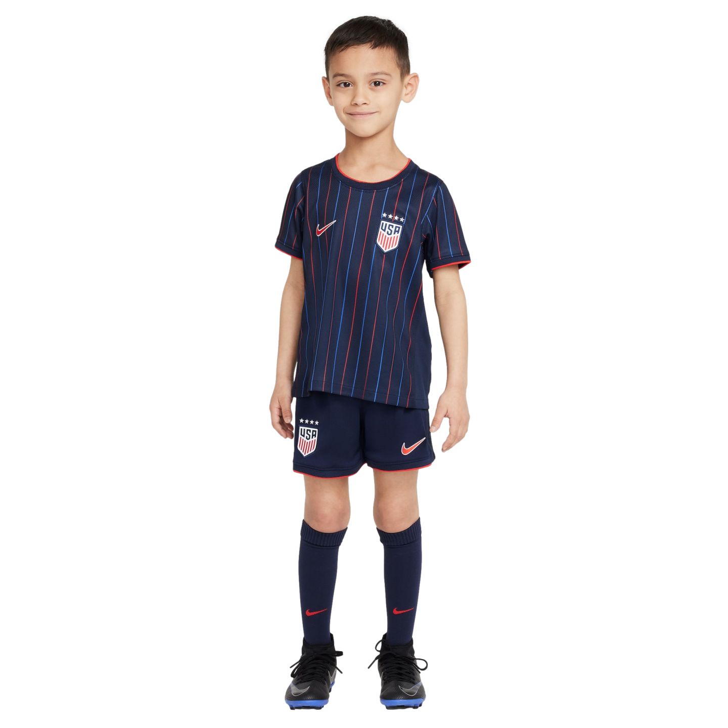 A young boy stands against a white background wearing a dark blue USA soccer kit with red and blue vertical stripes, matching shorts and socks, and black soccer cleats.