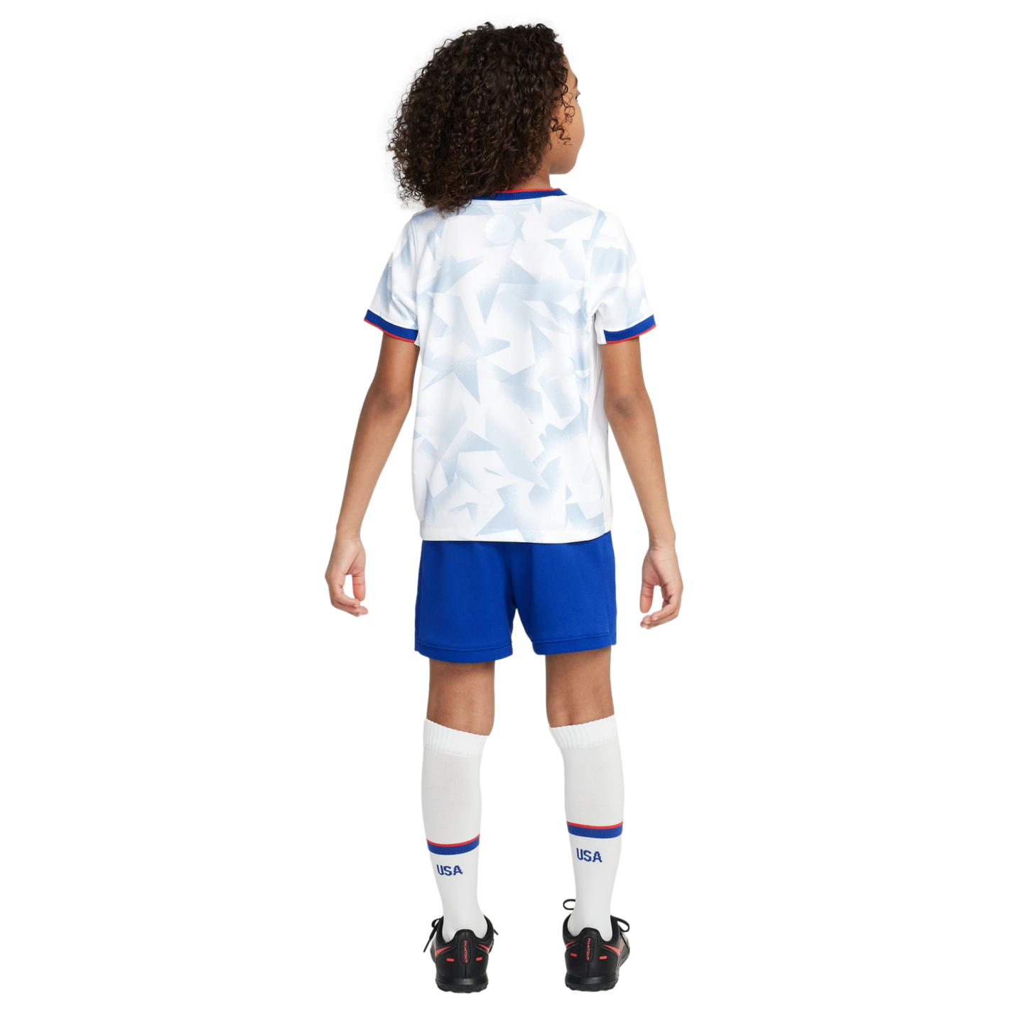 Back view of a child wearing a white USA soccer kit with geometric patterns, blue shorts, white socks with "USA" text, and black cleats.