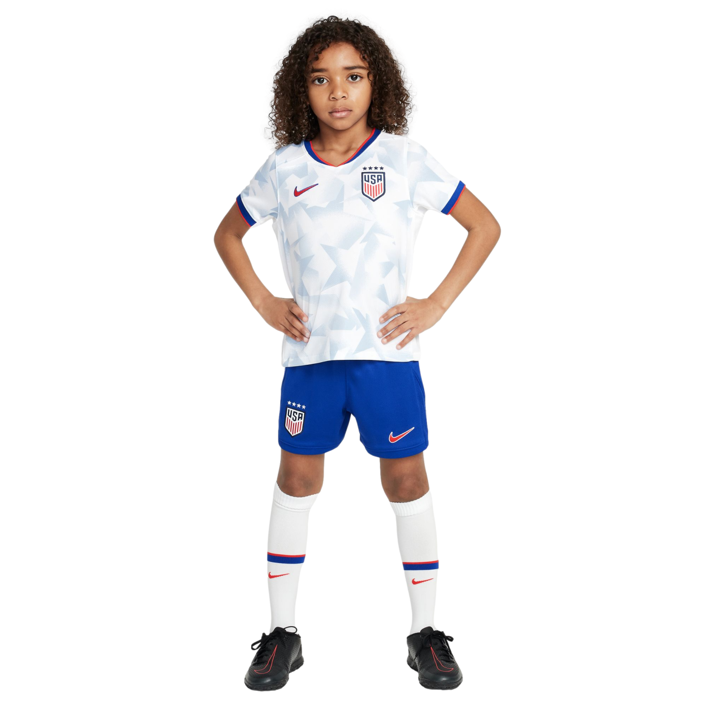 A young child in a white USA soccer uniform, blue shorts, and black cleats stands with hands on hips against a white background.