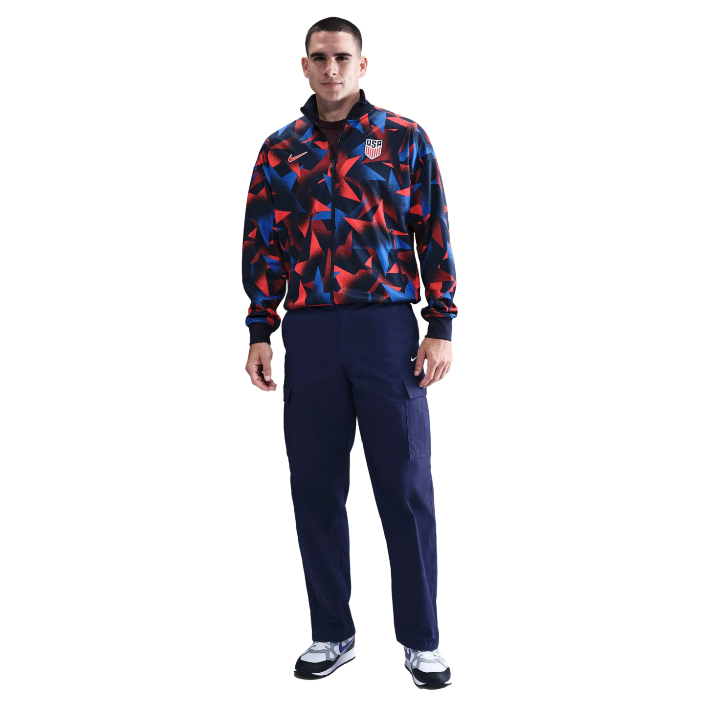 A man wears a red, blue, and black patterned Nike USA tracksuit jacket, navy cargo pants, and white sneakers against a white background.