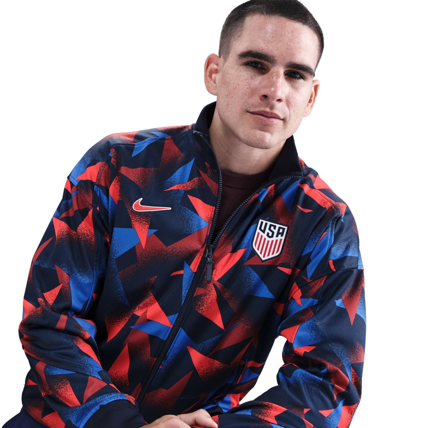 A man with freckles wears a red and blue geometric Nike USA national soccer team jacket against a white background.
