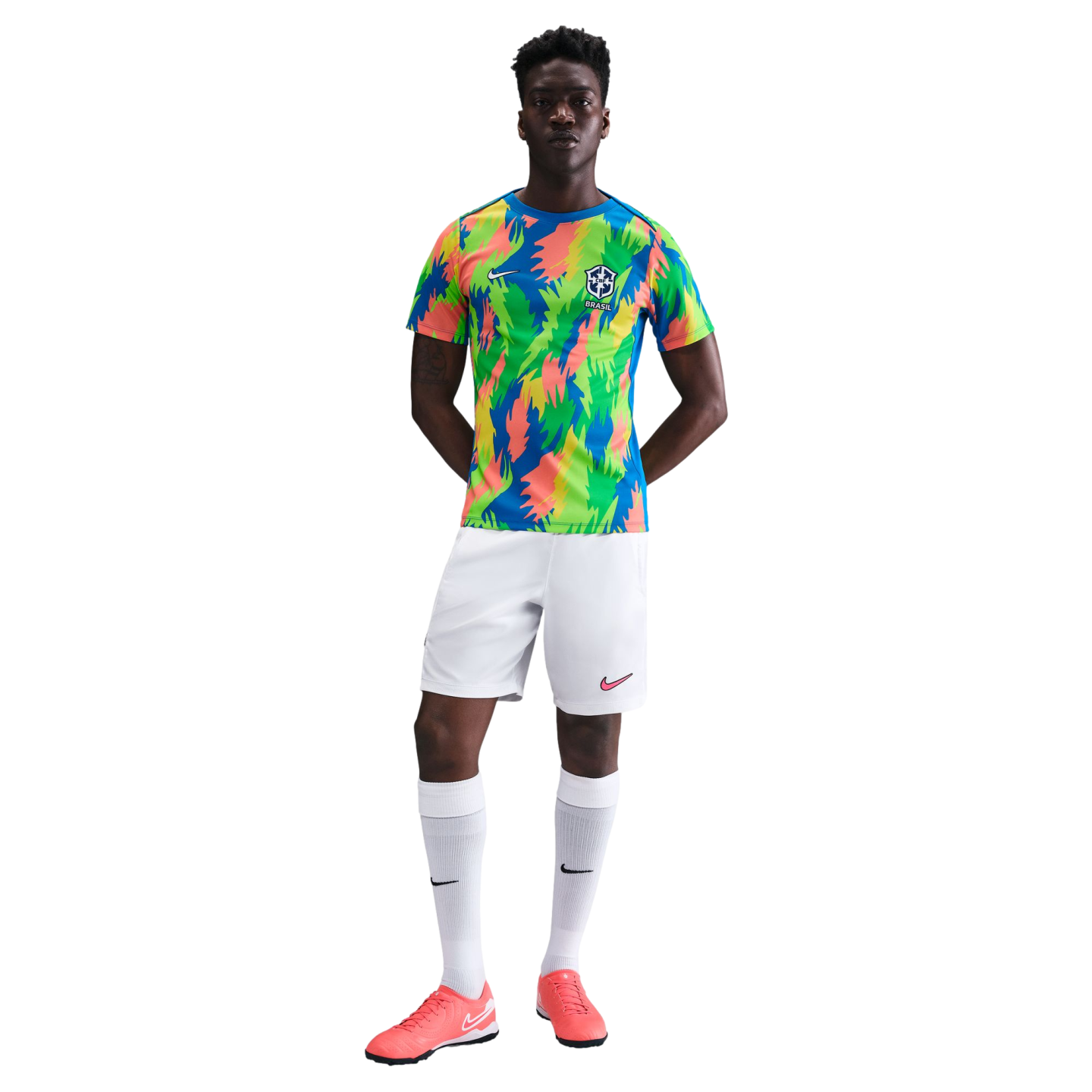 A Black man models a colorful Nike Brazil soccer kit with an abstract pattern jersey, white shorts and socks, and pink cleats against a white background.