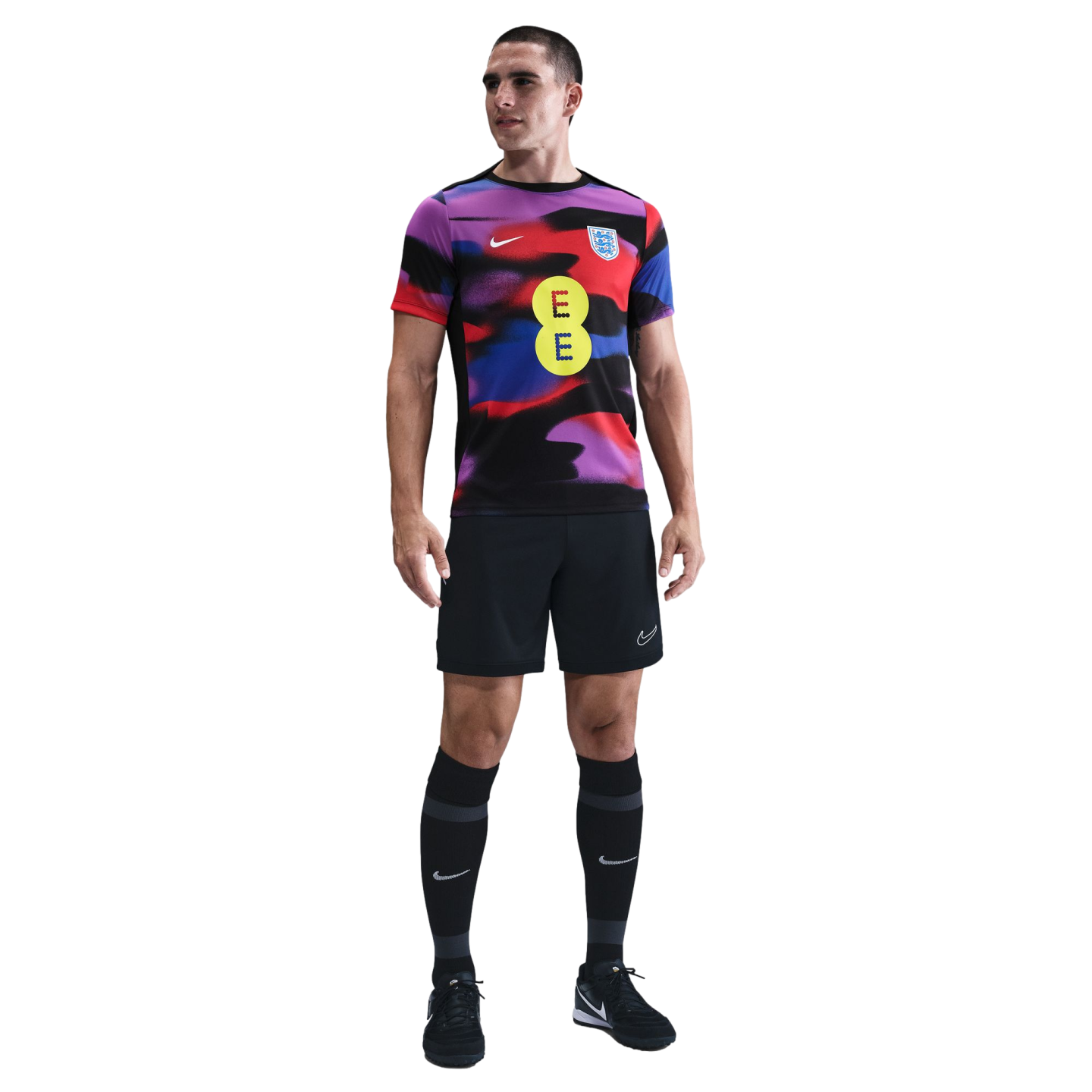 A man models a multicolored England football training kit with a yellow EE logo, black shorts, and black knee-high socks against a white background.