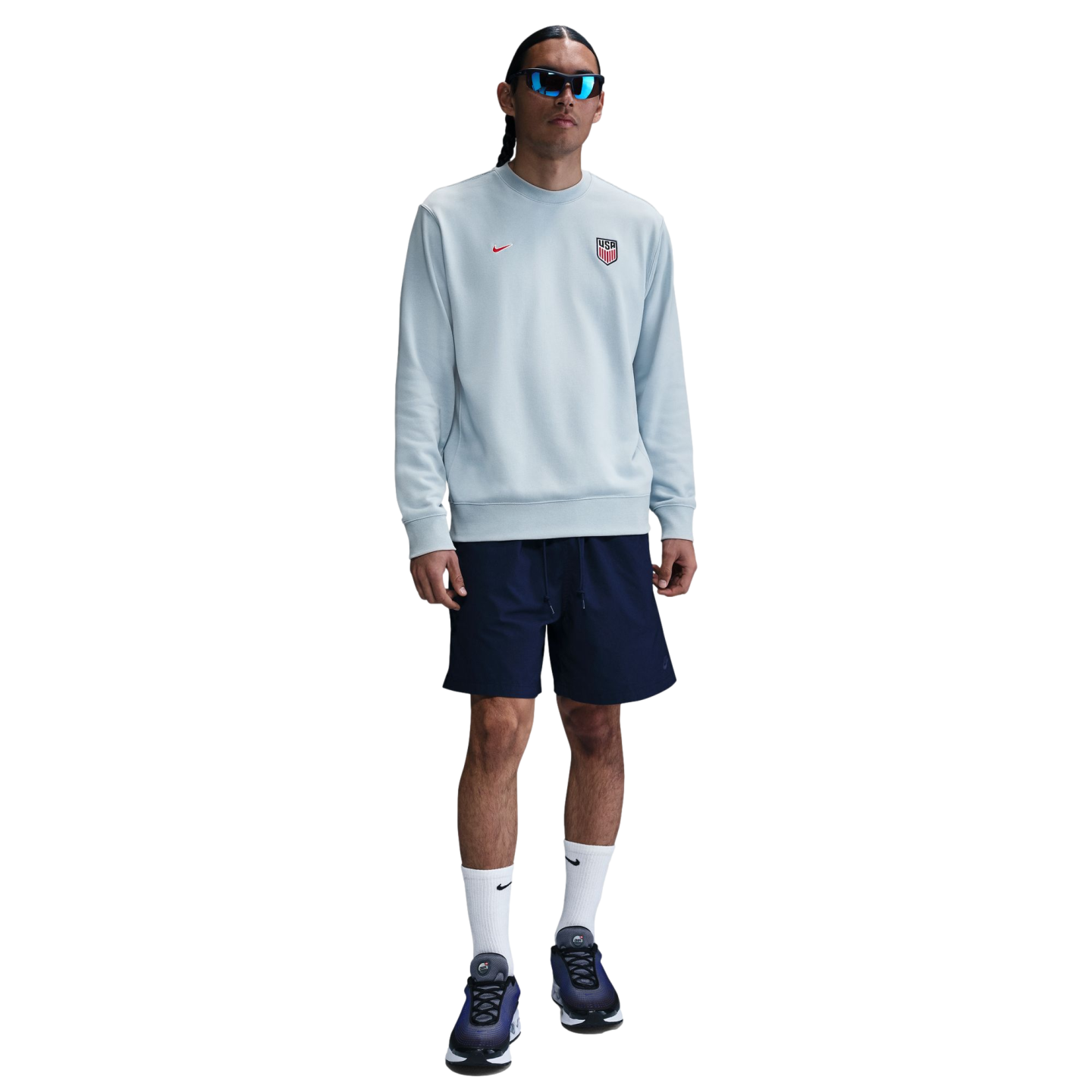 A man in a light blue Nike USA sweatshirt, navy shorts, white socks, and blue mirrored sunglasses against a white background.