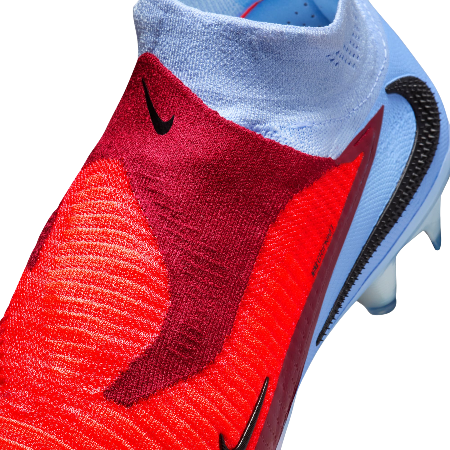 Close-up of a red and light blue Nike soccer boot featuring a textured Gripknit upper, black Nike swoosh, and a laceless ankle collar.