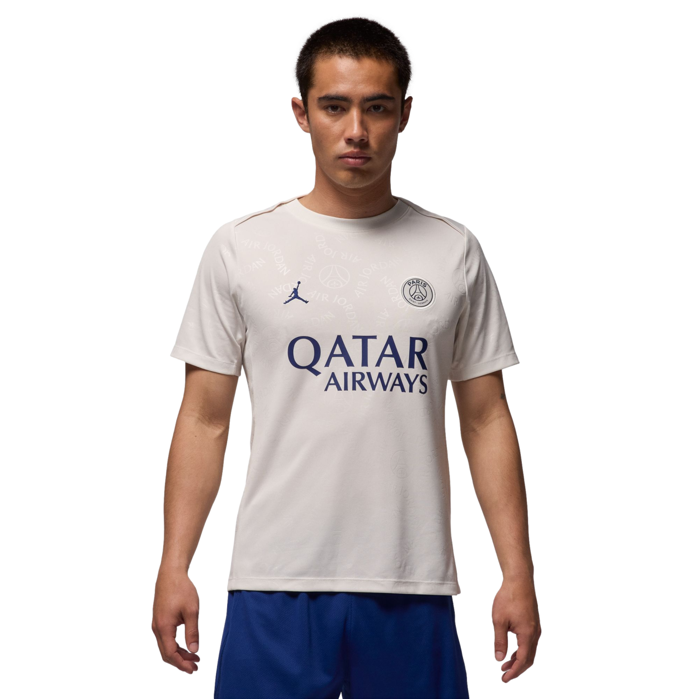 A male model wearing a cream-colored PSG x Jordan soccer jersey and blue shorts, standing against a white background.