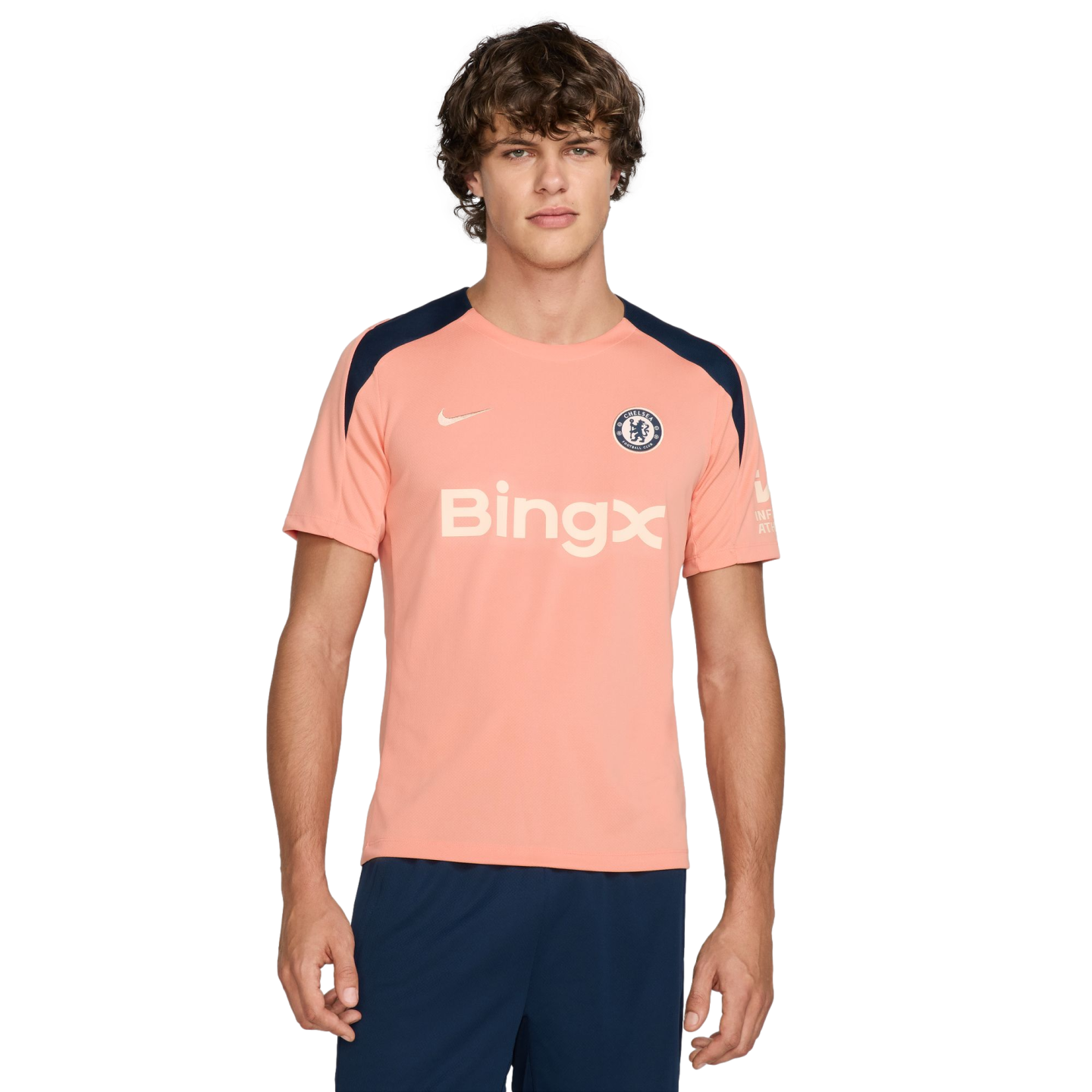 A young man with wavy brown hair wears a peach-colored Nike Chelsea FC training shirt with a "BingX" logo and navy blue shoulder panels, paired with navy shorts against a white background.