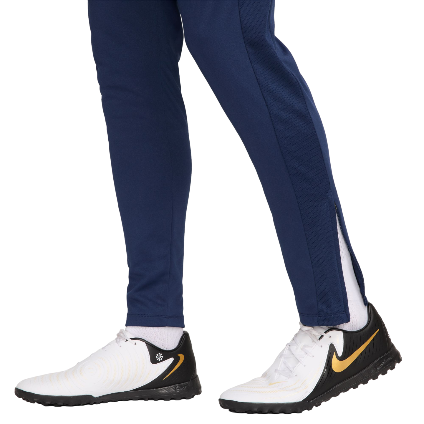 Side view of a person wearing navy blue track pants and white, black, and gold Nike turf shoes against a white background.