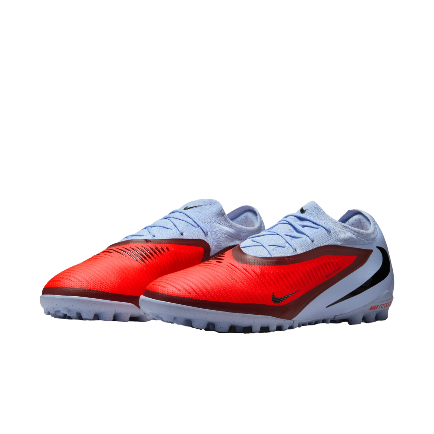 A pair of light blue and red Nike Phantom soccer turf shoes with black swoosh logos and grey outsoles, set against a black background.