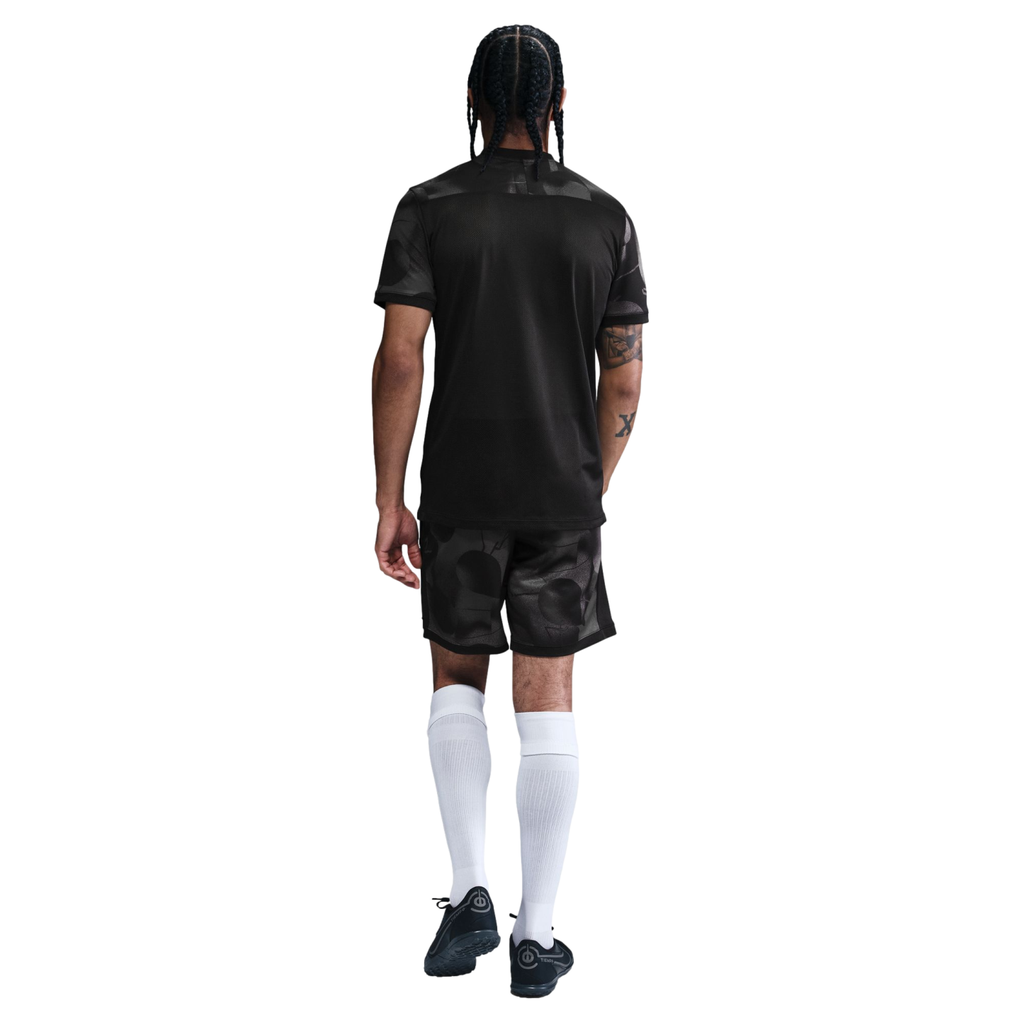 Back view of a man with braided hair wearing a black soccer jersey, patterned shorts, tall white socks, and black cleats.