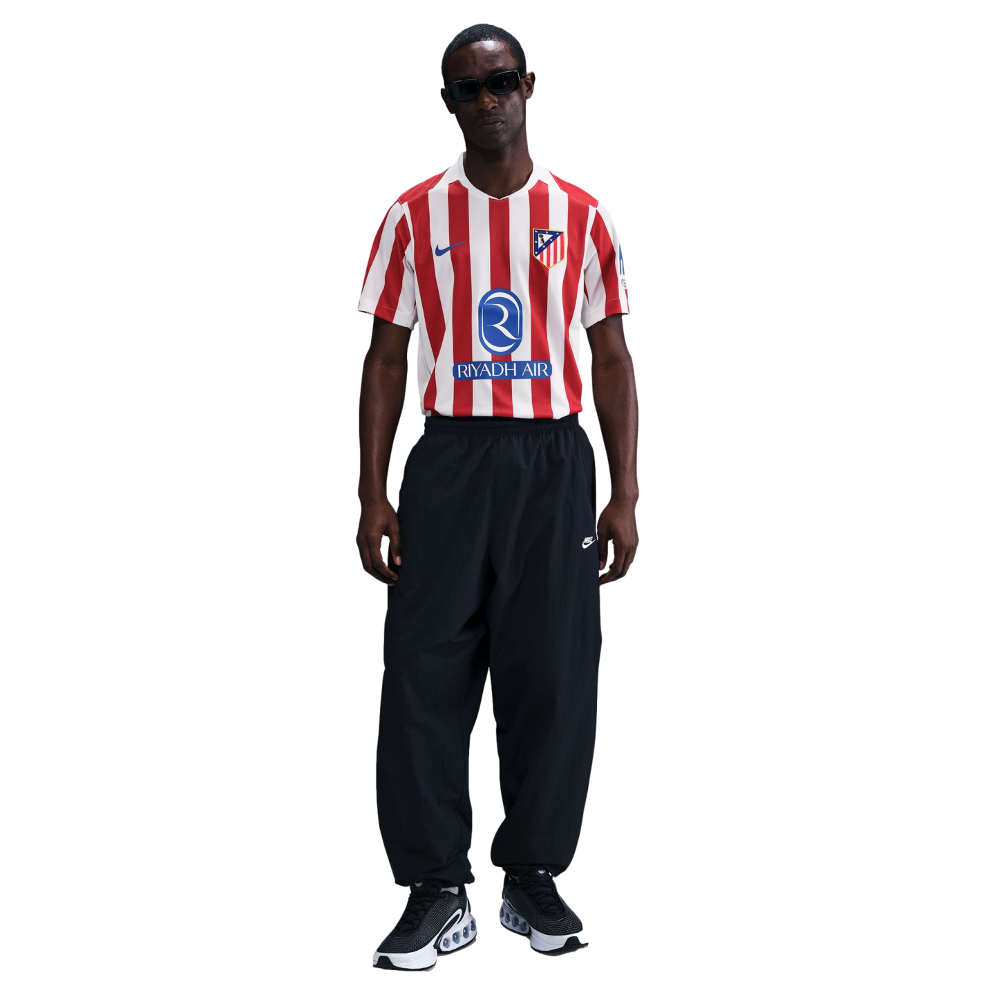 A man stands wearing a red and white striped Atlético de Madrid jersey, black Nike track pants, and black sunglasses against a white background.