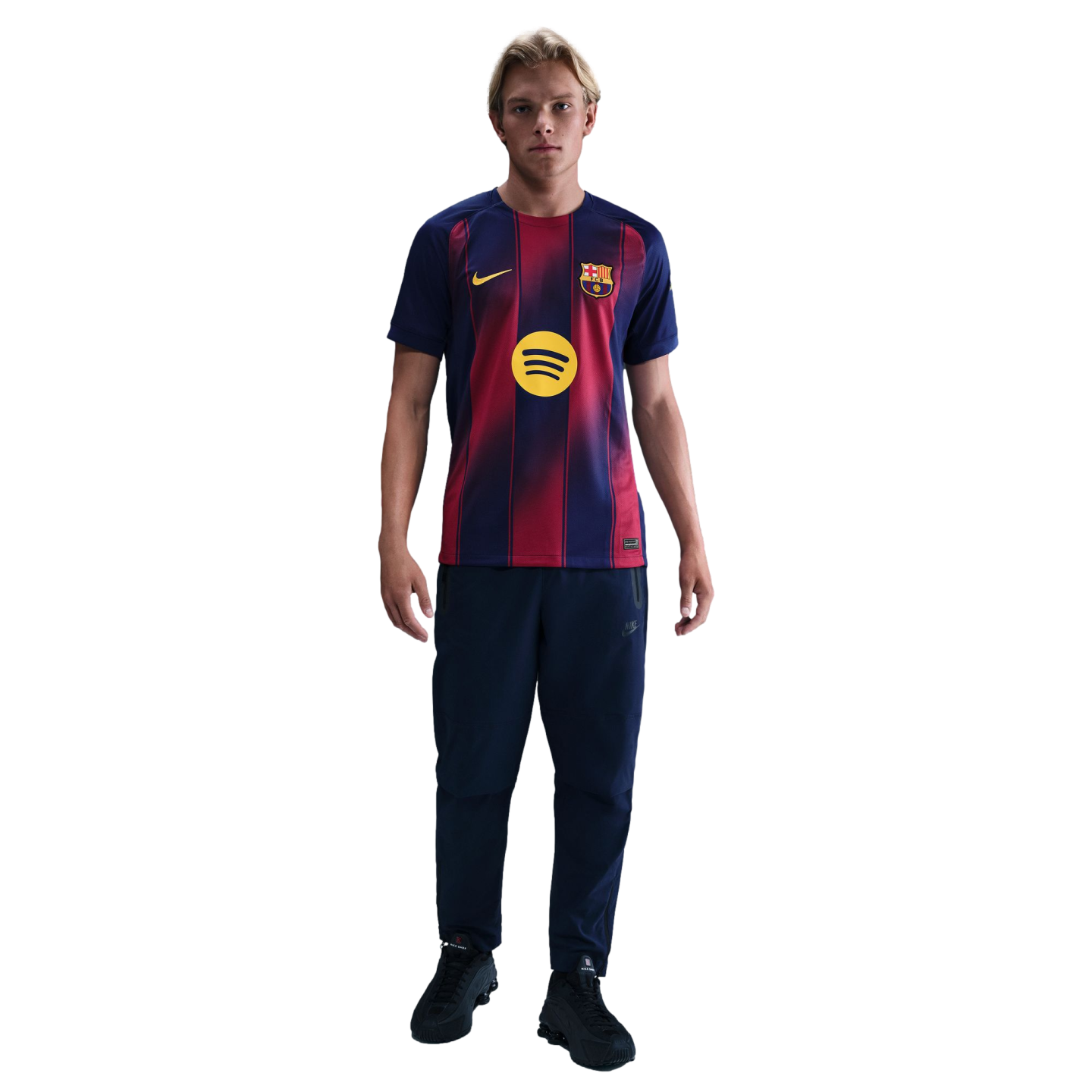 A blonde man wearing an FC Barcelona football jersey, navy track pants, and black trainers, standing against a solid black background.
