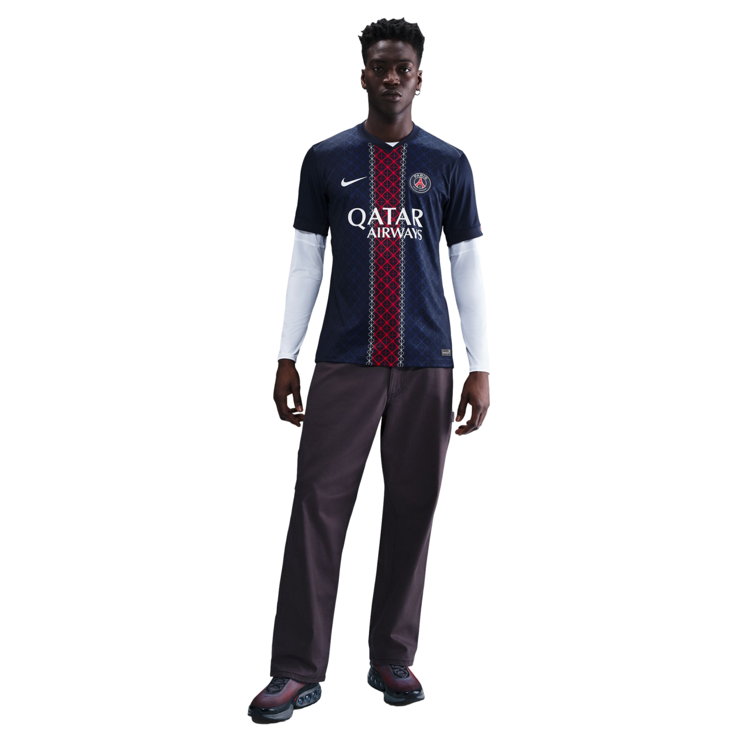 A man models a navy blue Paris Saint-Germain home jersey over a white long-sleeve shirt, dark trousers, and sneakers against a plain white background.