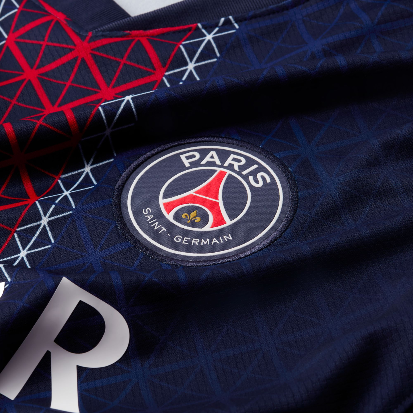 Close-up of a navy blue Paris Saint-Germain football jersey with a geometric pattern, the club’s circular logo patch, and a partial white "R" at the bottom.