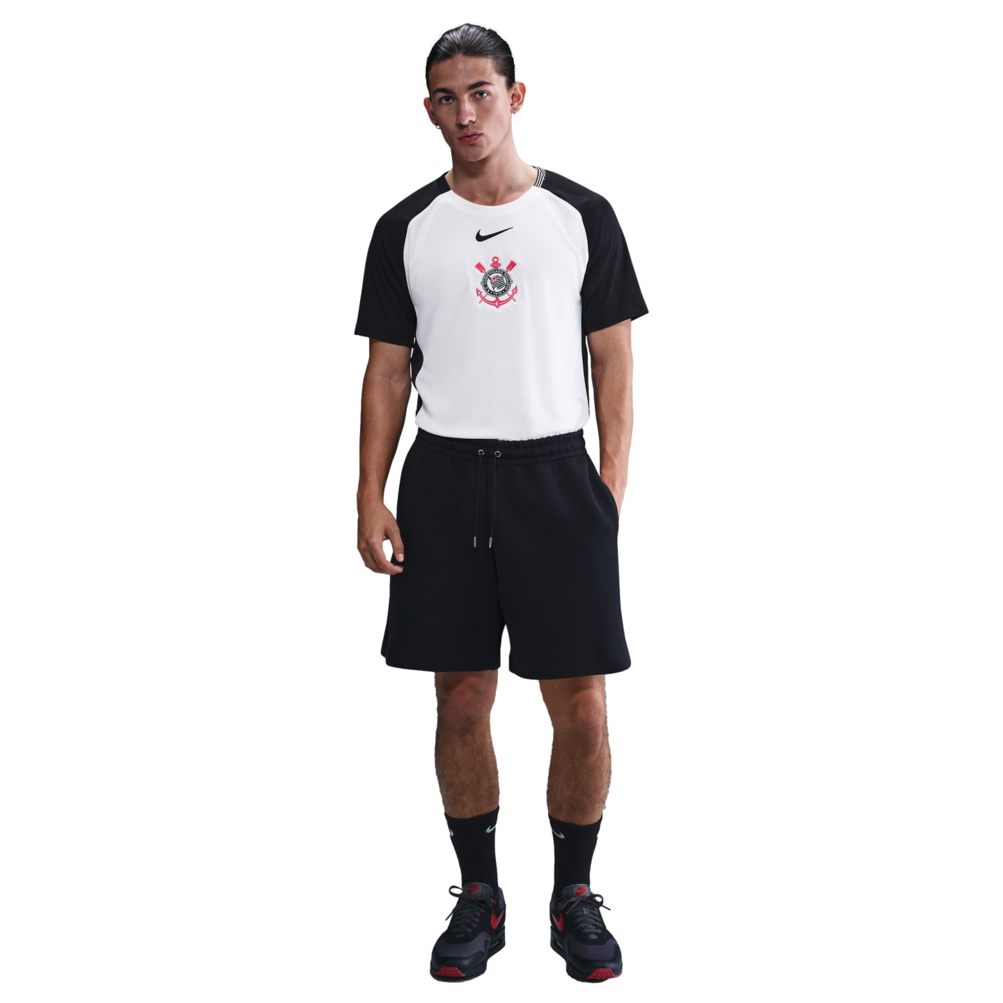 A man wearing a Nike Sport Club Corinthians Paulista t-shirt, black athletic shorts, black crew socks, and black sneakers, standing against a white background.