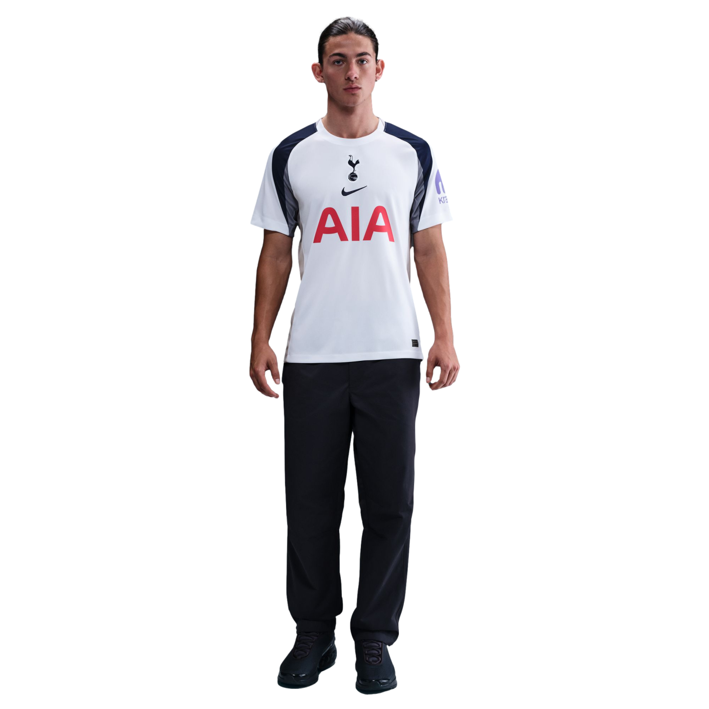 A man stands against a white background wearing a white and navy Tottenham Hotspur football shirt, black trousers, and black trainers.
