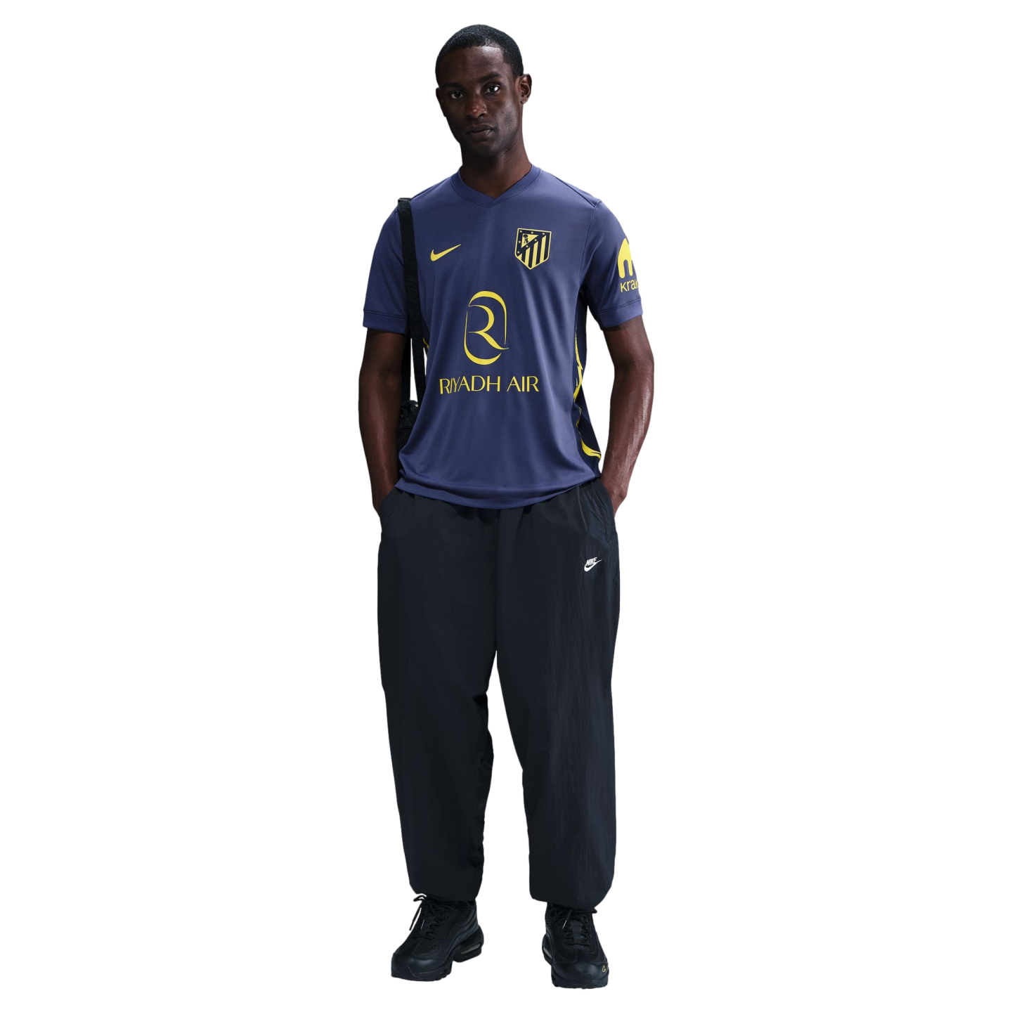 A man standing in a blue and yellow Atlético Madrid soccer jersey, black trousers, and black sneakers.