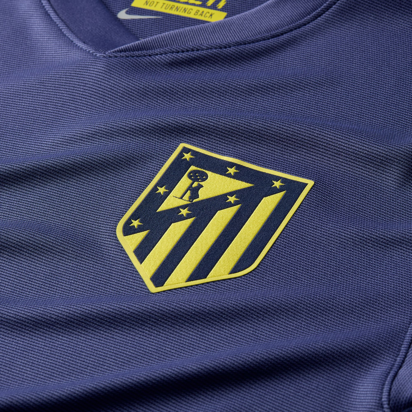 Close-up of a navy blue Atletico Madrid jersey with a yellow and blue team crest. The textured mesh fabric includes a Nike collar label that reads "NOT TURNING BACK."
