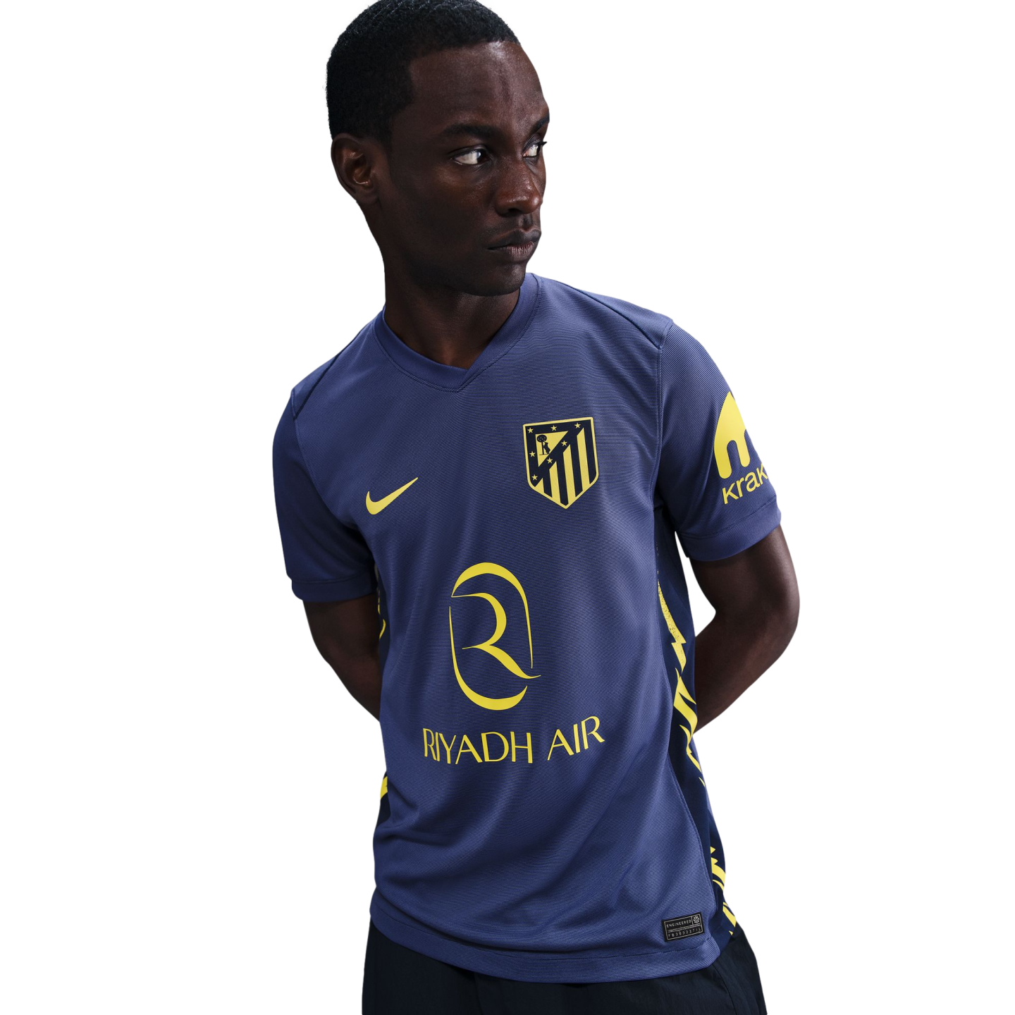 A man wearing a blue Atlético Madrid soccer jersey with yellow Nike and Riyadh Air logos, looking off to the side against a plain white background.