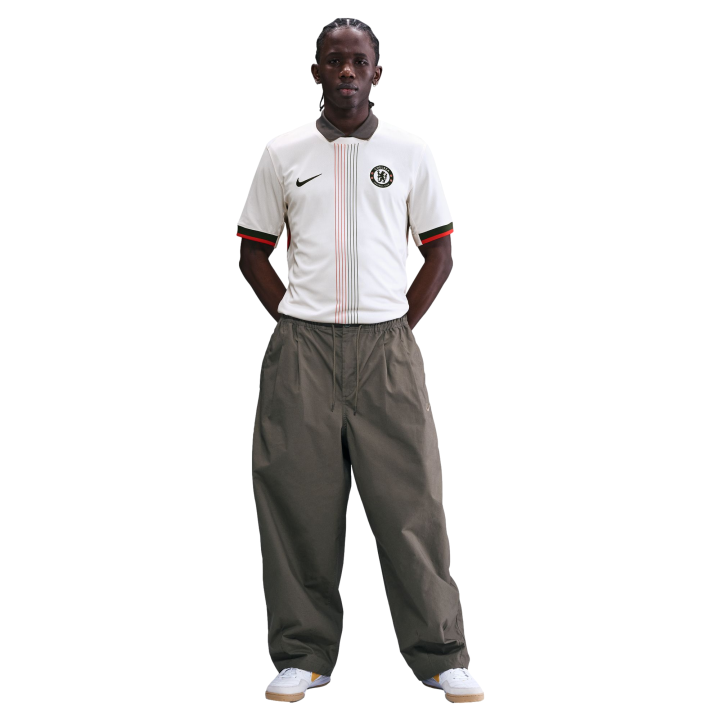 A man in a white Nike Chelsea FC polo shirt, loose olive green trousers, and white sneakers, standing against a white background with his hands behind his back.
