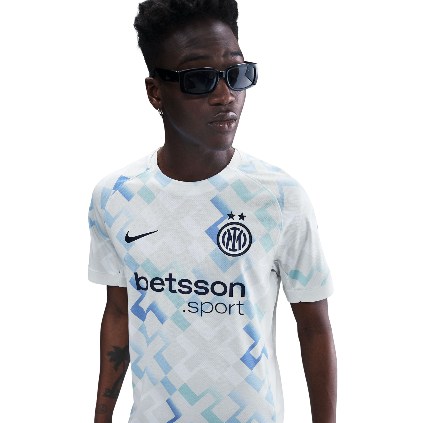 A young Black man in sunglasses wears an Inter Milan football jersey with a blue and teal geometric pattern, set against a solid white background.