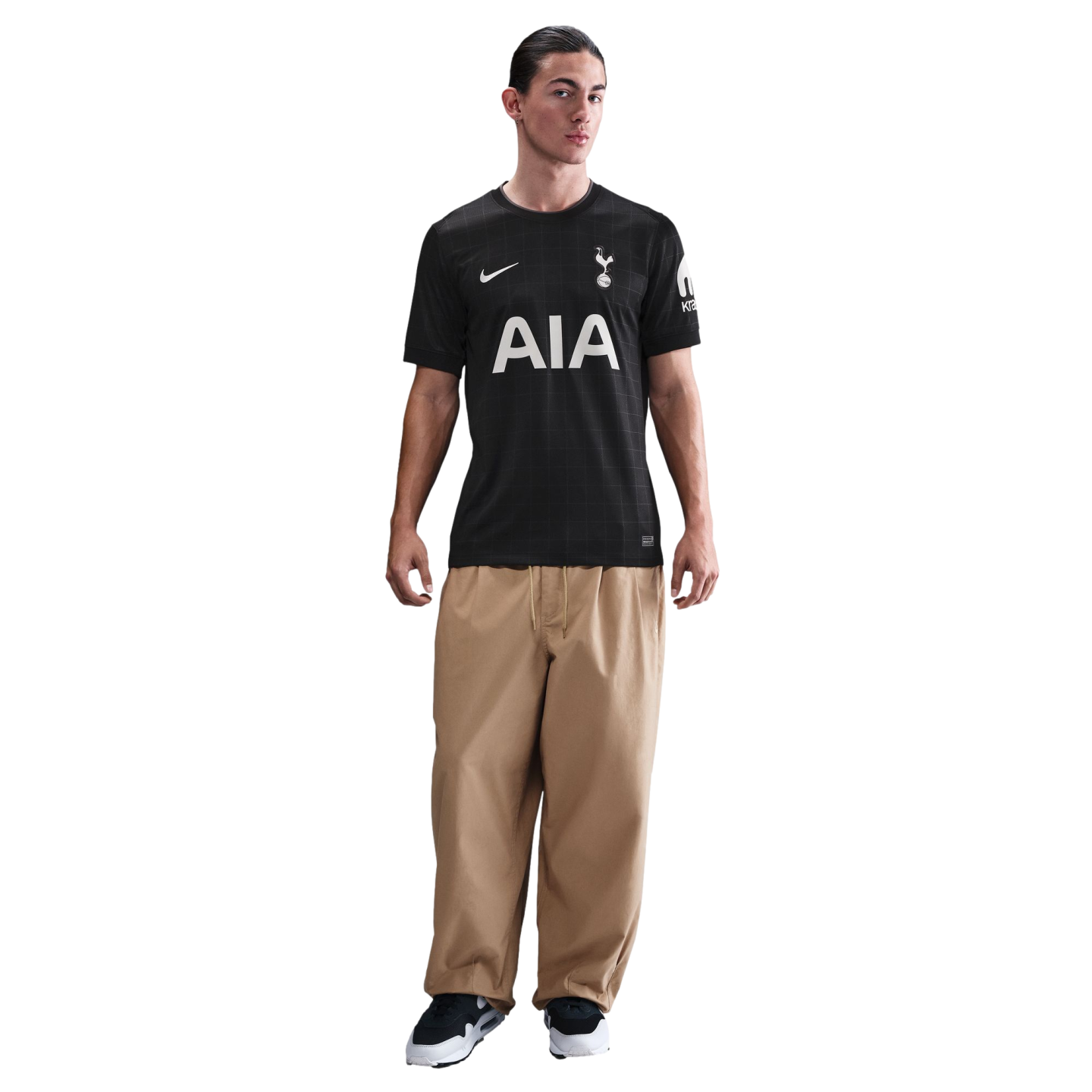 A man wearing a black Tottenham Hotspur soccer jersey and baggy khaki trousers stands against a black background.