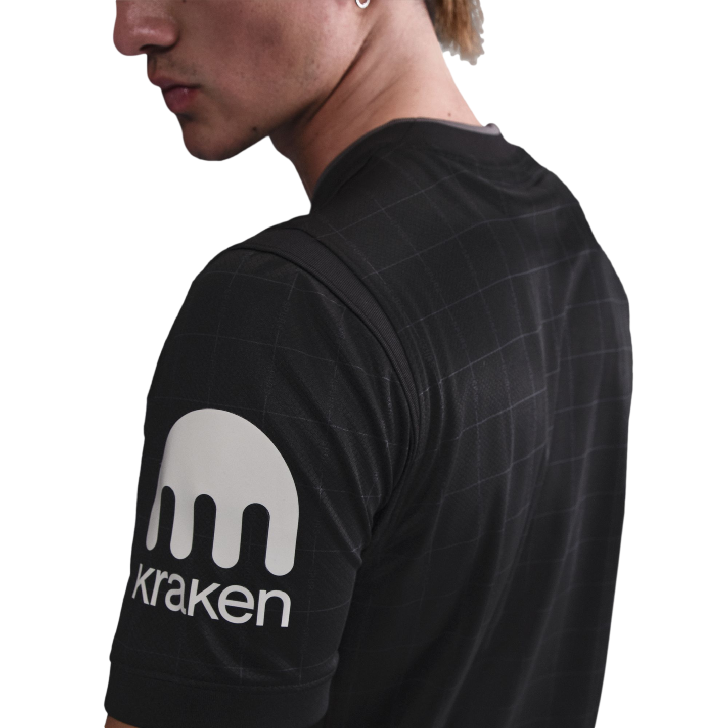 Close-up of a person wearing a black grid-patterned shirt with a large white Kraken logo and text on the sleeve.