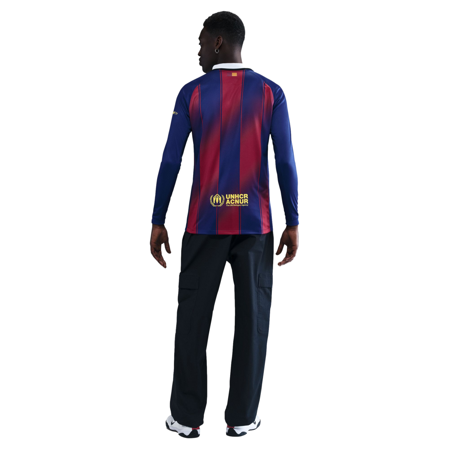 Back view of a person wearing a long-sleeved blue and red striped soccer jersey, dark cargo pants, and white sneakers, against a solid black background.