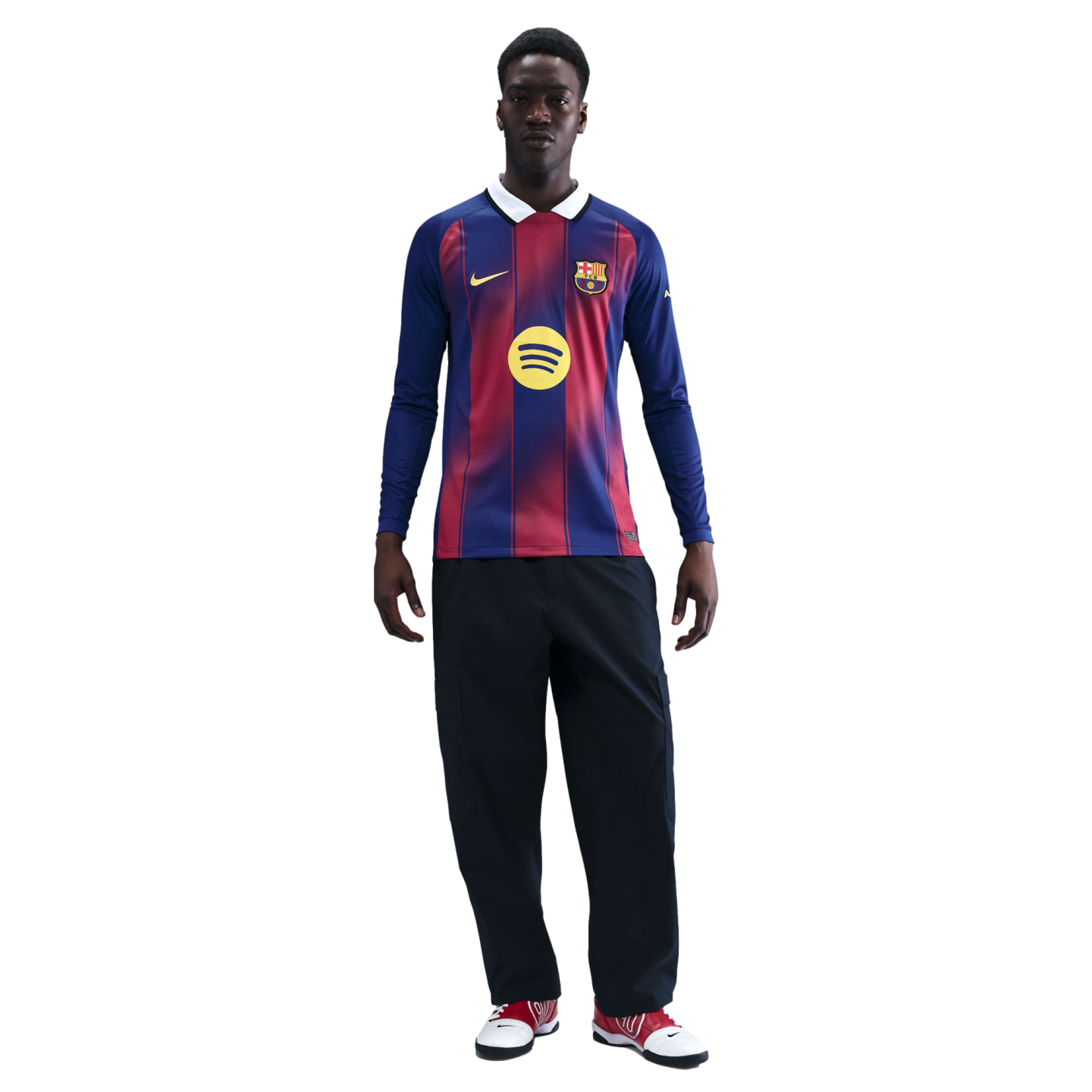 A man stands against a black background wearing a long-sleeved FC Barcelona jersey, dark pants, and red and white sneakers.