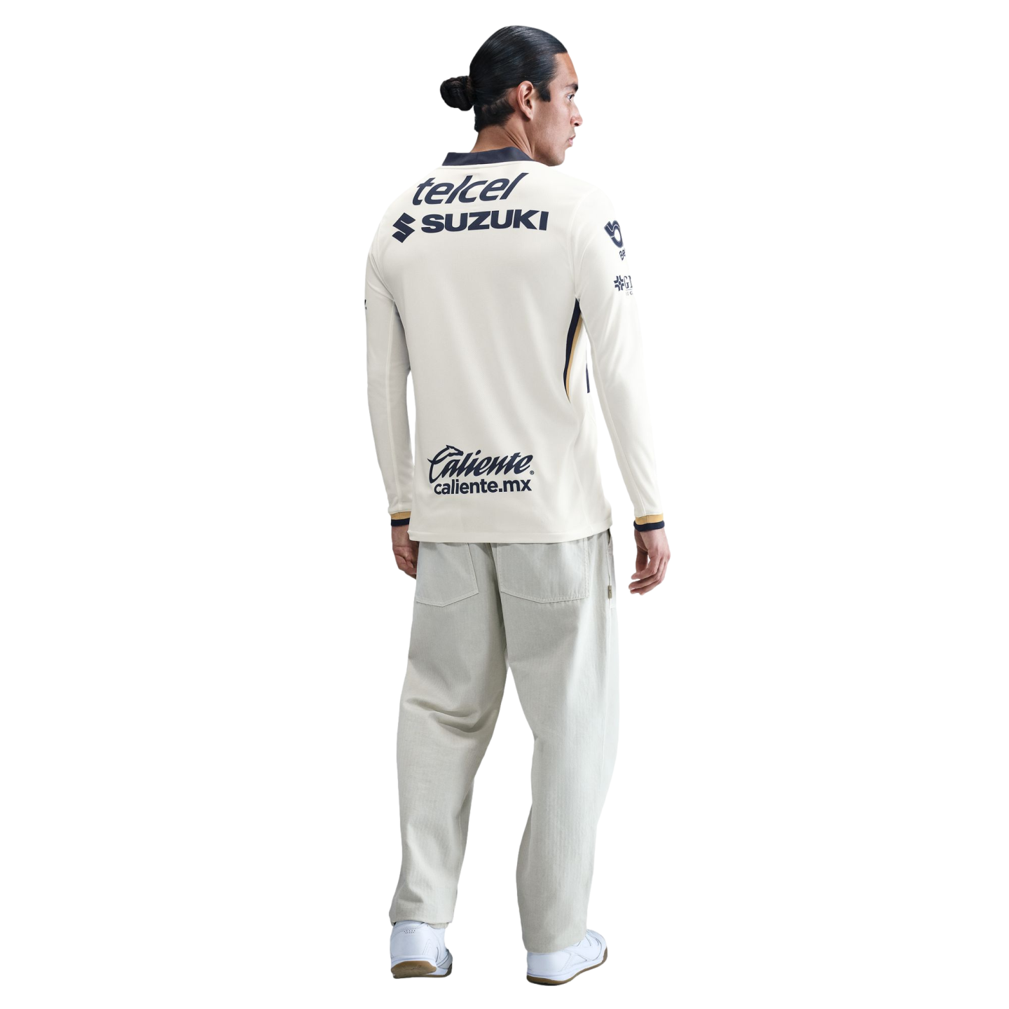 Back view of a man wearing a cream long-sleeved soccer jersey with sponsor logos and light gray wide-leg pants against a white background.