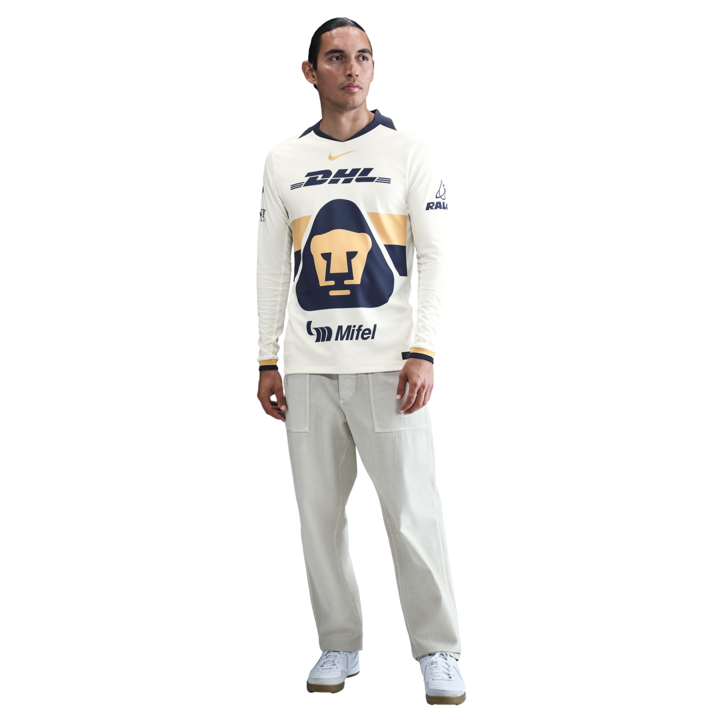 Man wearing a long-sleeved cream Pumas UNAM soccer jersey, light gray trousers, and white sneakers, standing against a plain white background.