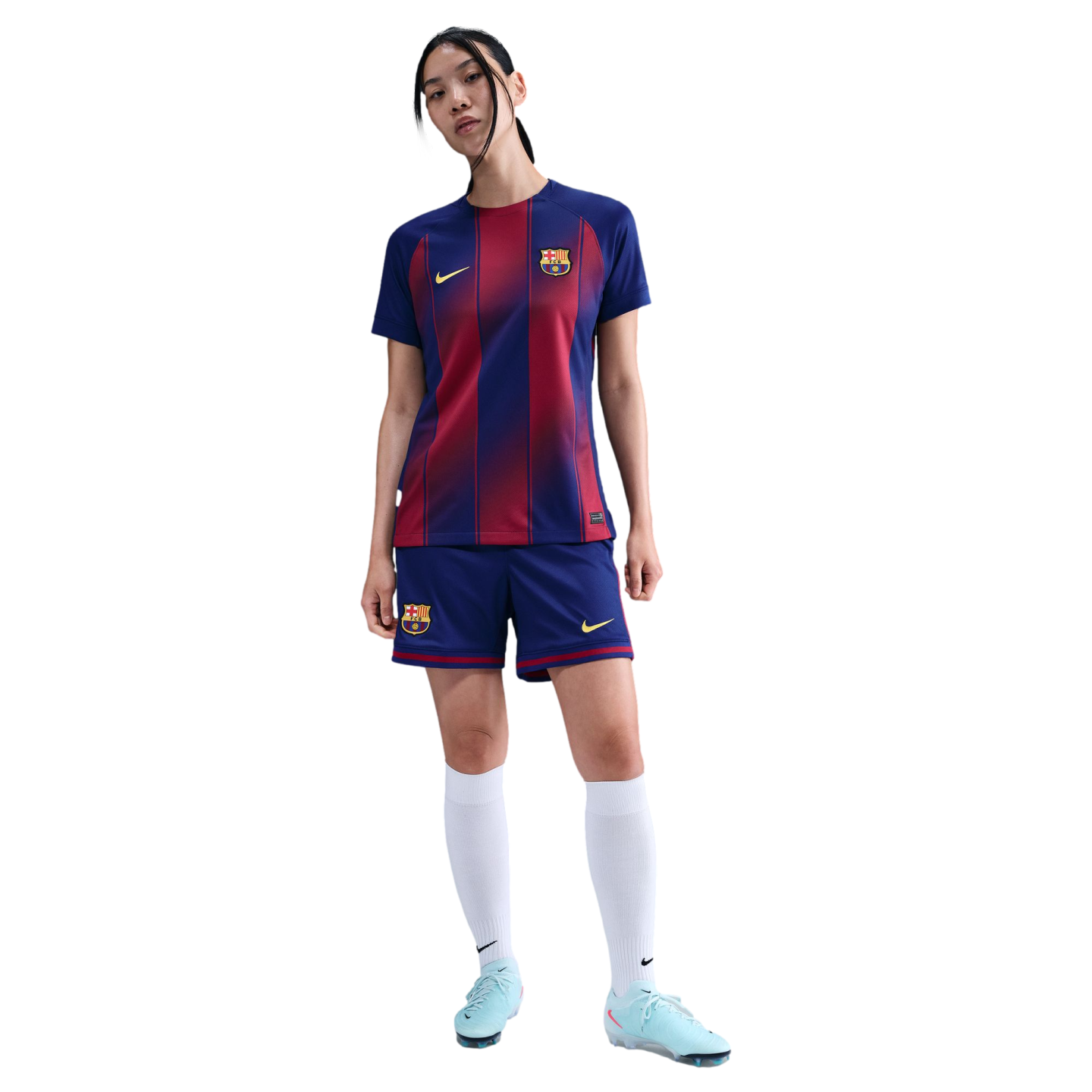 A woman wears an FC Barcelona soccer kit with a blue and red striped jersey, blue shorts, white socks, and light blue cleats, standing against a black background.