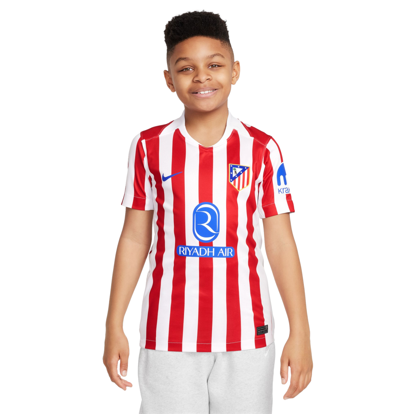 A smiling young boy wearing a red and white striped Atlético Madrid soccer jersey and light grey shorts, standing against a plain white background.