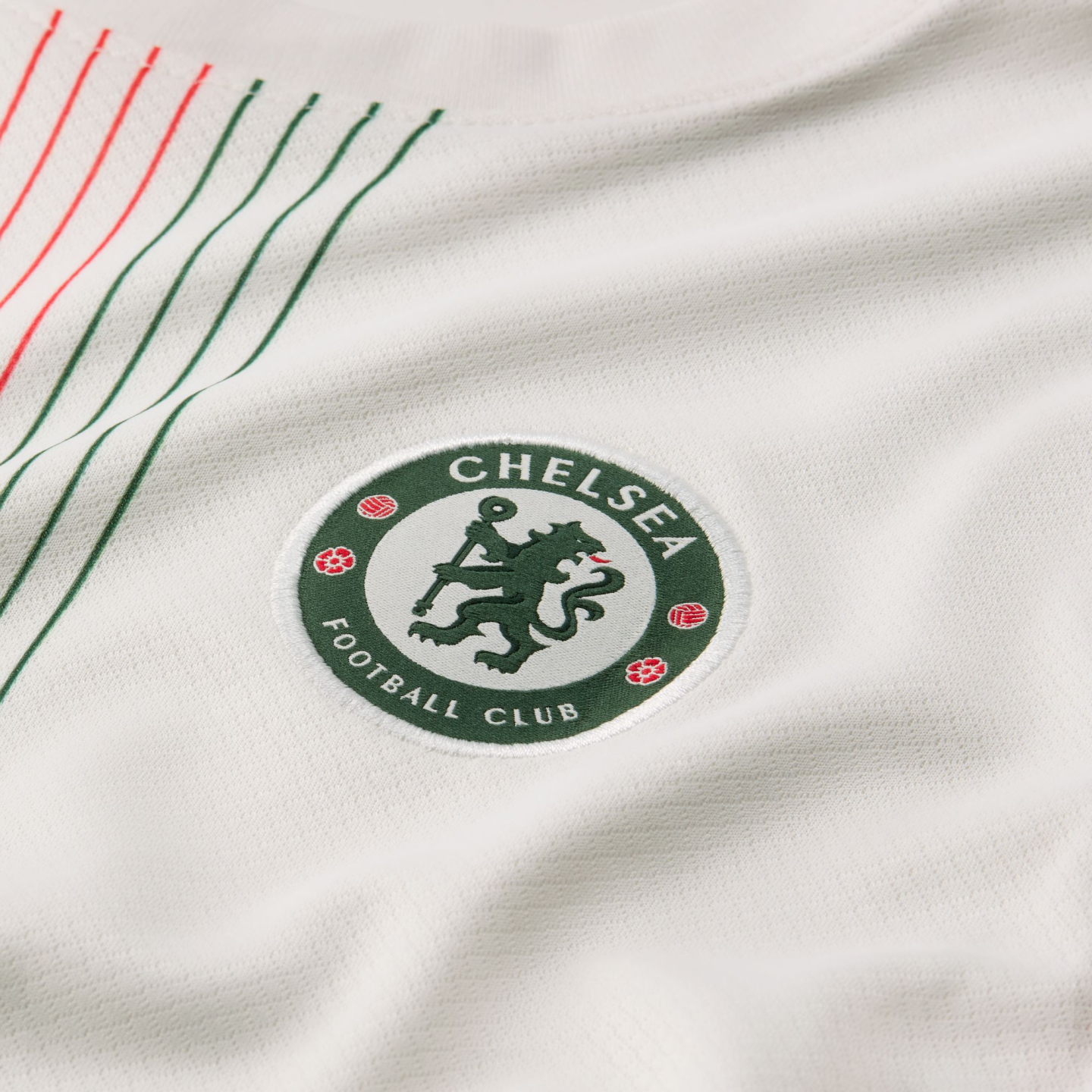 Close-up of a green Chelsea Football Club crest on a white textured jersey with thin red and green diagonal stripes in the corner.