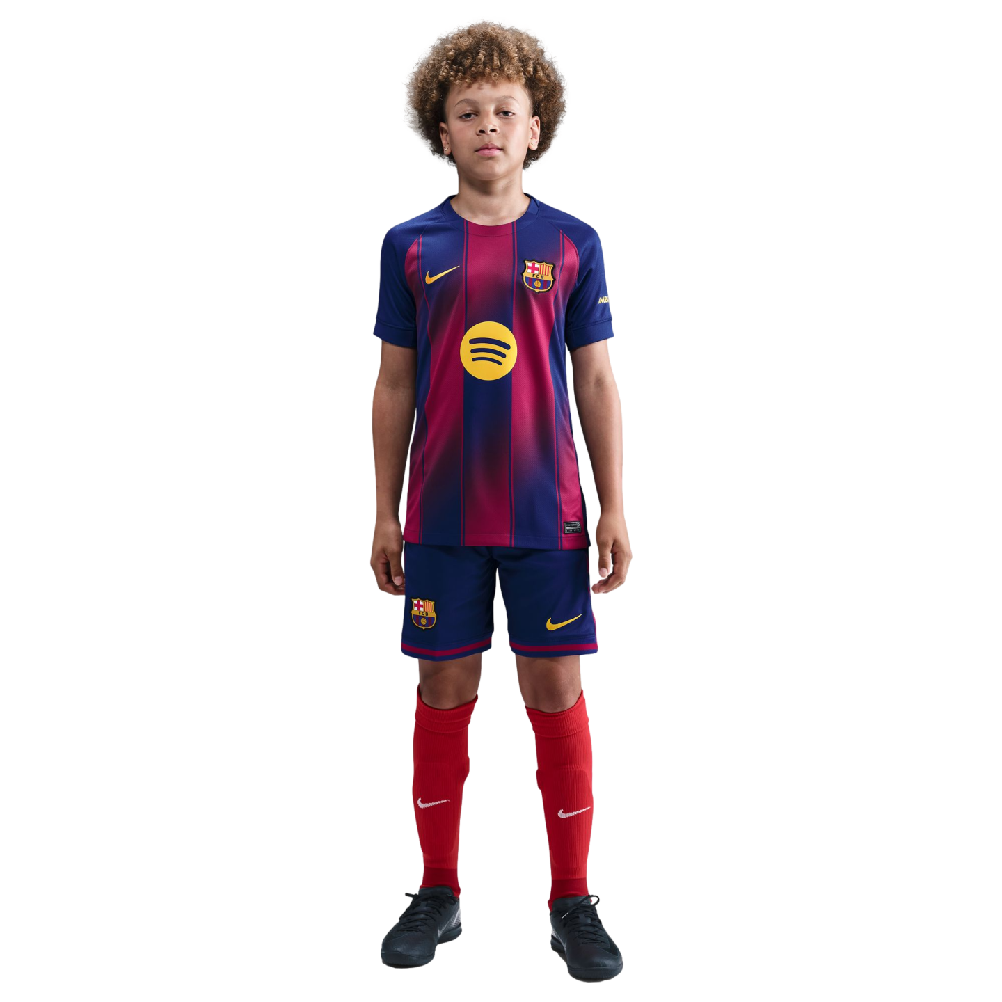 A young boy with curly hair stands against a black background wearing a full FC Barcelona football kit: blue and red striped jersey, blue shorts, and red socks.