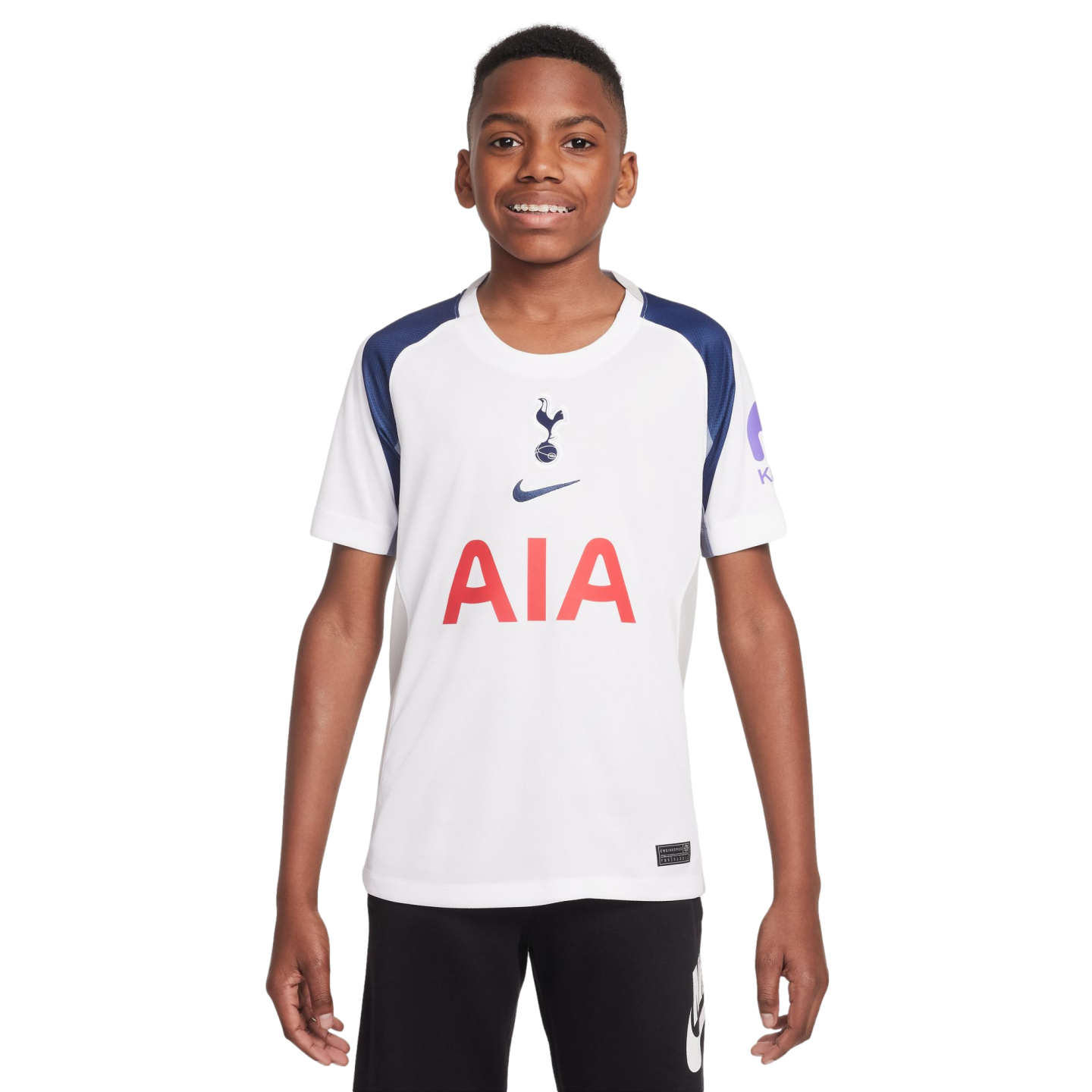 A smiling boy wears a white and navy Tottenham Hotspur soccer jersey with the red AIA logo and black shorts, standing against a white background.