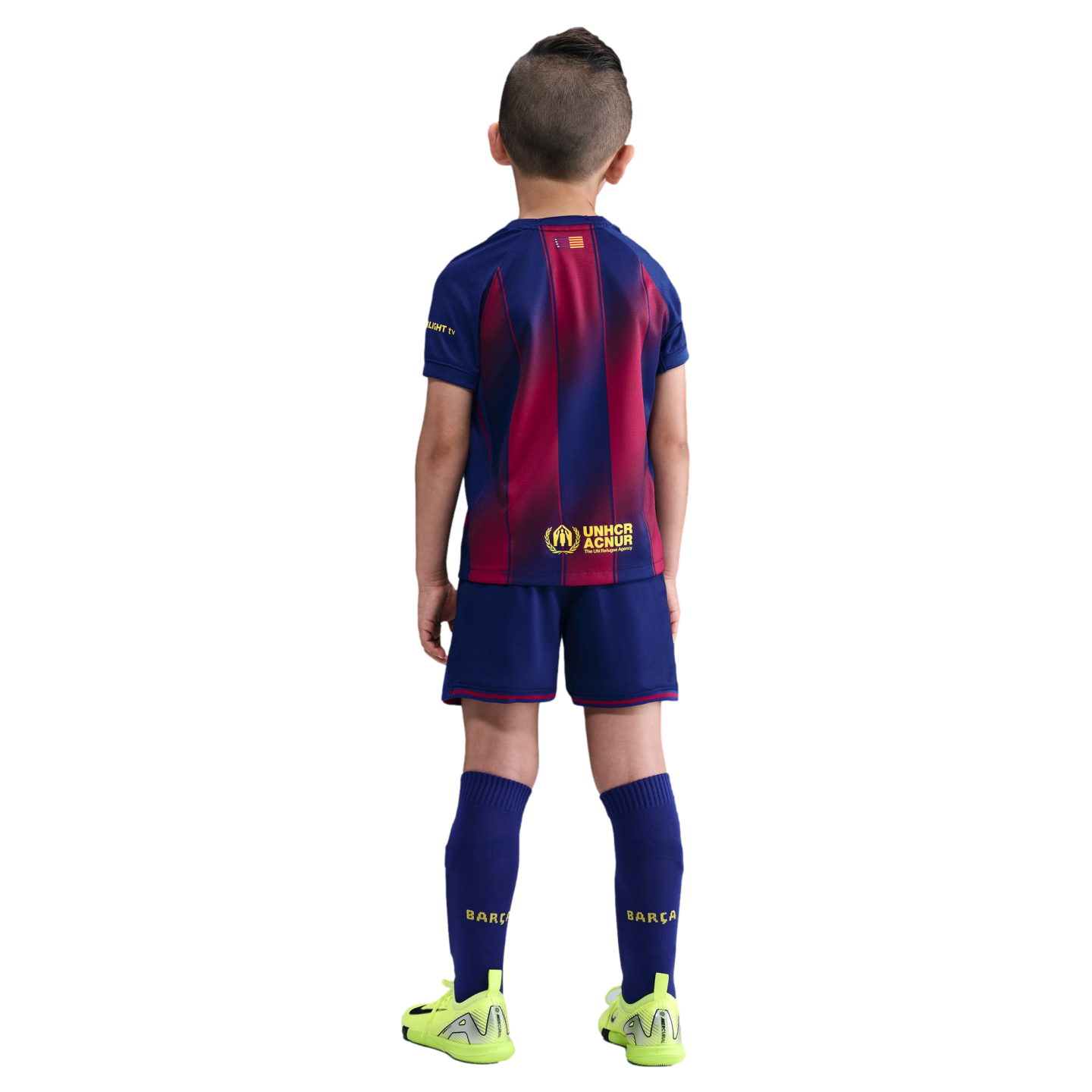 Back view of a boy in a blue and maroon FC Barcelona football kit and neon green cleats against a solid black background.